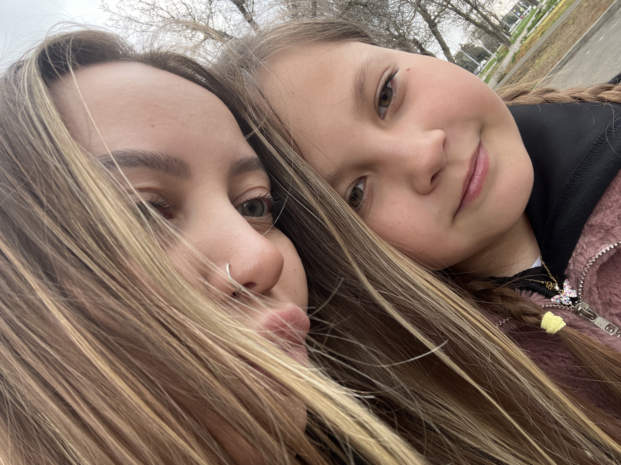 Two females, presumably a young adult and a child, are closely posed for a selfie. The adult is positioned on the left, while the child is on the right. The adult has light brown hair with a visible nose ring. The child has braided hair, and a butterfly necklace is visible. In the background, trees and a section of a building are visible, suggesting an urban environment. The photo was taken outdoors in Novopokrovskaya, Russia.