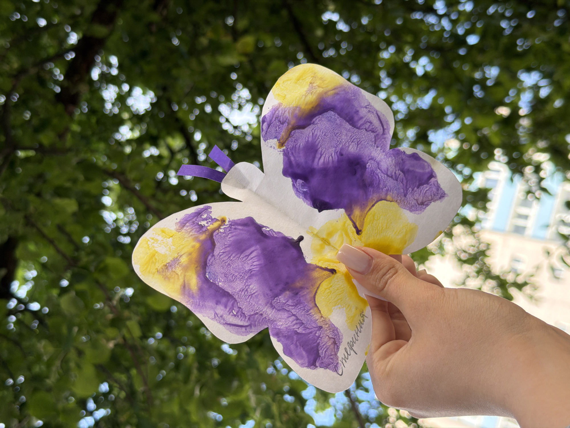A Caucasian female hand holds a paper butterfly painted with purple and yellow watercolors. The butterfly has a purple ribbon attached to its center. Part of a handwritten signature, "Стефания," is visible on the butterfly. The background shows green trees with light bokeh, a white building, and blue sky. This photo was taken in Chaikovsky, Russia.