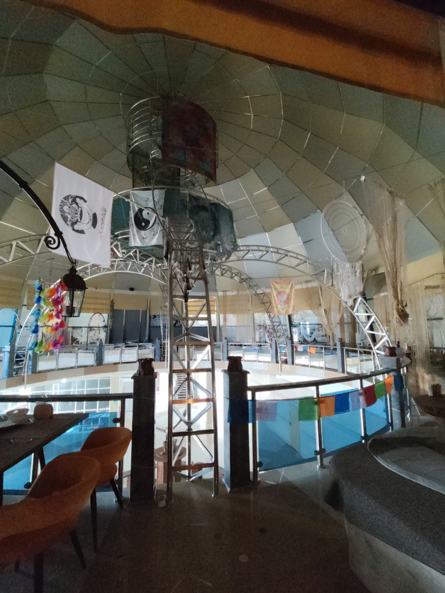 This image depicts the interior of a large, multi-level building. The architecture features a distinctive geodesic dome ceiling with metallic structural supports visible throughout. Various decorative elements are present, including flags with symbols such as yin and yang, a lantern with colorful feathers hanging from it, and prayer flags strung along railings. Several levels of balconies or walkways are visible, with railings and some furniture, including chairs and a table. The overall aesthetic suggests a communal or spiritual space, possibly a center for meditation or art. The scene appears to be unoccupied by people. While the provided information states the location is Sochi, Russia, there are no specific visual cues within the image that definitively identify it as such.