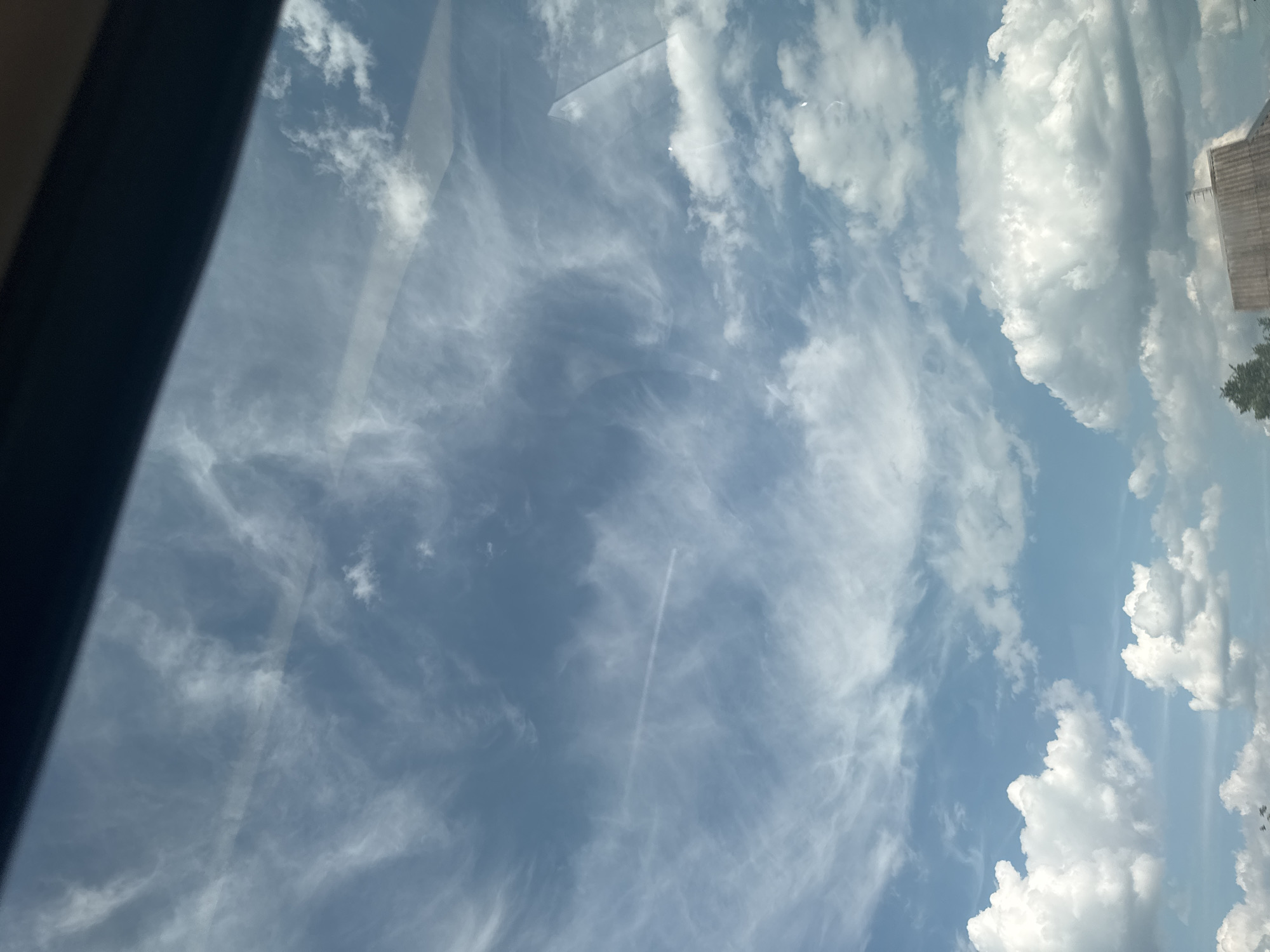 The image displays a daytime view of the sky over Markivka, Ukraine, captured through a reflective surface, likely a window or windshield, indicated by subtle glare and reflections. The upper and middle portions of the sky are dominated by light blue hues, traversed by widespread, wispy cirrus clouds forming intricate, feathery patterns. A distinct, linear contrail, indicative of an aircraft's passage, extends horizontally across the central-upper sky. Towards the bottom, several bright white, puffy cumulus clouds are present. The lower-most section of the image reveals elements of the ground, including a dark, sloped, corrugated roof structure on the right and a portion of dark green foliage, possibly from a coniferous tree, on the left. A dark, solid, diagonal obstruction is visible across the top of the frame. No individuals, specific actions, or direct interactions are discernible within the frame.