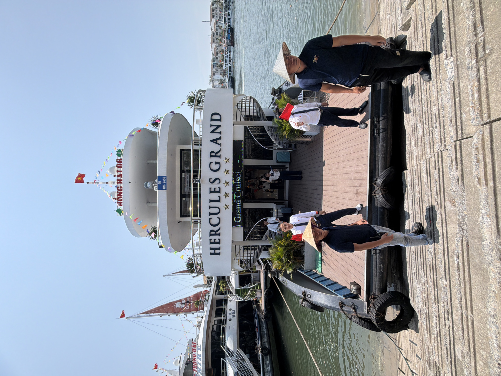 The image captures a bustling dockside scene in Tuan Chau Ward, Vietnam, under a clear, bright blue sky. A large white multi-deck cruise ship, named "HERCULES GRAND," is docked, adorned with colorful pennant banners. Behind it, another boat with a red sail and a Vietnamese flag is partially visible, showing text like "PHONG HAI" and "QN 99..." on its hull.
Several people are present. On the wooden deck of the "HERCULES GRAND," one individual stands wearing a traditional Vietnamese conical hat (nón lá), a dark blue polo shirt, and light grey pants. Nearby, another person in a white long-sleeved shirt, black trousers, and a red bag over their shoulder also appears to wear a conical hat. On the concrete pier, a third person in a dark polo shirt and black trousers is visible. Inside the ship's entrance, additional people, possibly crew members, can be seen.
The cruise ship is securely moored to a concrete pier with ropes and black tire fenders. The bright sun casts strong shadows on the textured concrete, indicating clear and sunny weather.
Visible text on the main ship includes "HERCULES GRAND" in large letters, and below it, "Grand Cruise" with a star rating. A blue sign near the entrance displays "99 QN-9456," likely a registration number. Partial text "HONG HA R" is also visible on the ship's side. The presence of conical hats and Vietnamese flags clearly situates the scene in Vietnam, depicting activity around a cruise embarkation or disembarkation point.