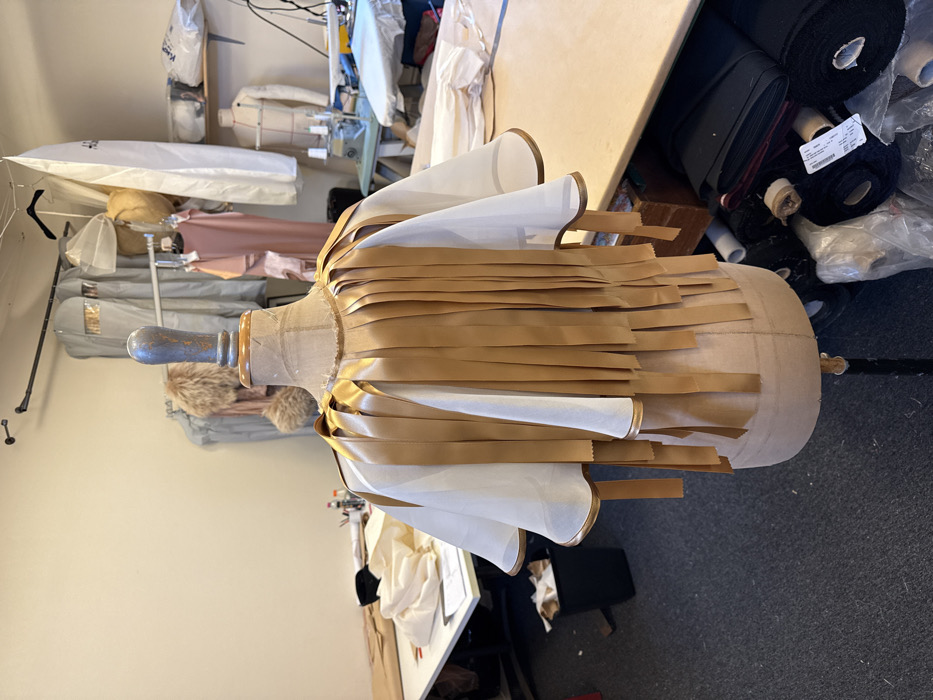 An indoor fashion design studio in Arcueil, France, is depicted. The central subject is a beige, fabric-covered dress form, elevated on a black metal stand with a light-colored wooden neck. A partially assembled garment adorns the dress form. This garment consists of a translucent white, short-sleeved cape-like layer with gold-piped edges, overlaid by multiple vertical strips of metallic gold ribbon. These ribbons, varying in length, are attached from the neckline, extending down past the waist, creating a fringed texture.
To the left, a light-colored work table contains various off-white fabric pieces, paper patterns, and a small container of writing instruments. A black waste bin, holding crumpled light brown material, rests beneath this table. The floor is covered with dark grey carpeting.
The cream-colored wall in the background features a metal garment rack. Hanging garments include items in clear plastic garment bags, a light pink dress, and a beige fur stole. A white garment bag with visible text is also suspended. Further to the right, a second light-colored table holds a light blue industrial sewing machine and an ironing board with an iron. A white male torso dress form stands in the background on this side. Rolls of dark-colored fabric are stored beneath the right table. The area is uniformly illuminated by overhead artificial lighting.