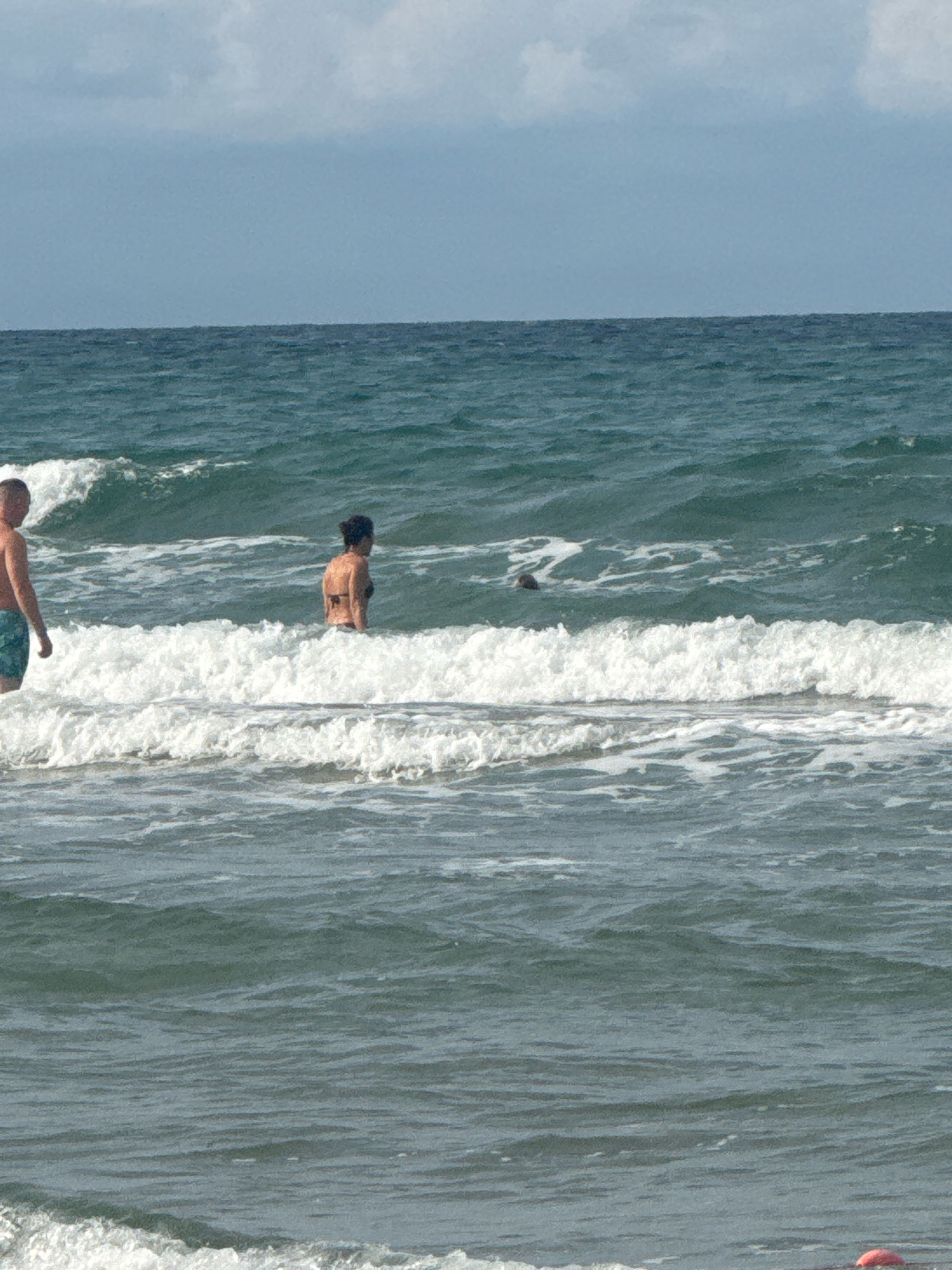 This is a medium shot, outdoor, eye-level photograph taken of the sea in Anapa, Russia. Two people are visible in the water. On the left, a man wearing green patterned swim trunks is facing away from the camera. On the right, a woman in a dark bikini is facing sideways, also away from the camera. The water is wavy with white foam from breaking waves. The sky is overcast with light blue and gray clouds.