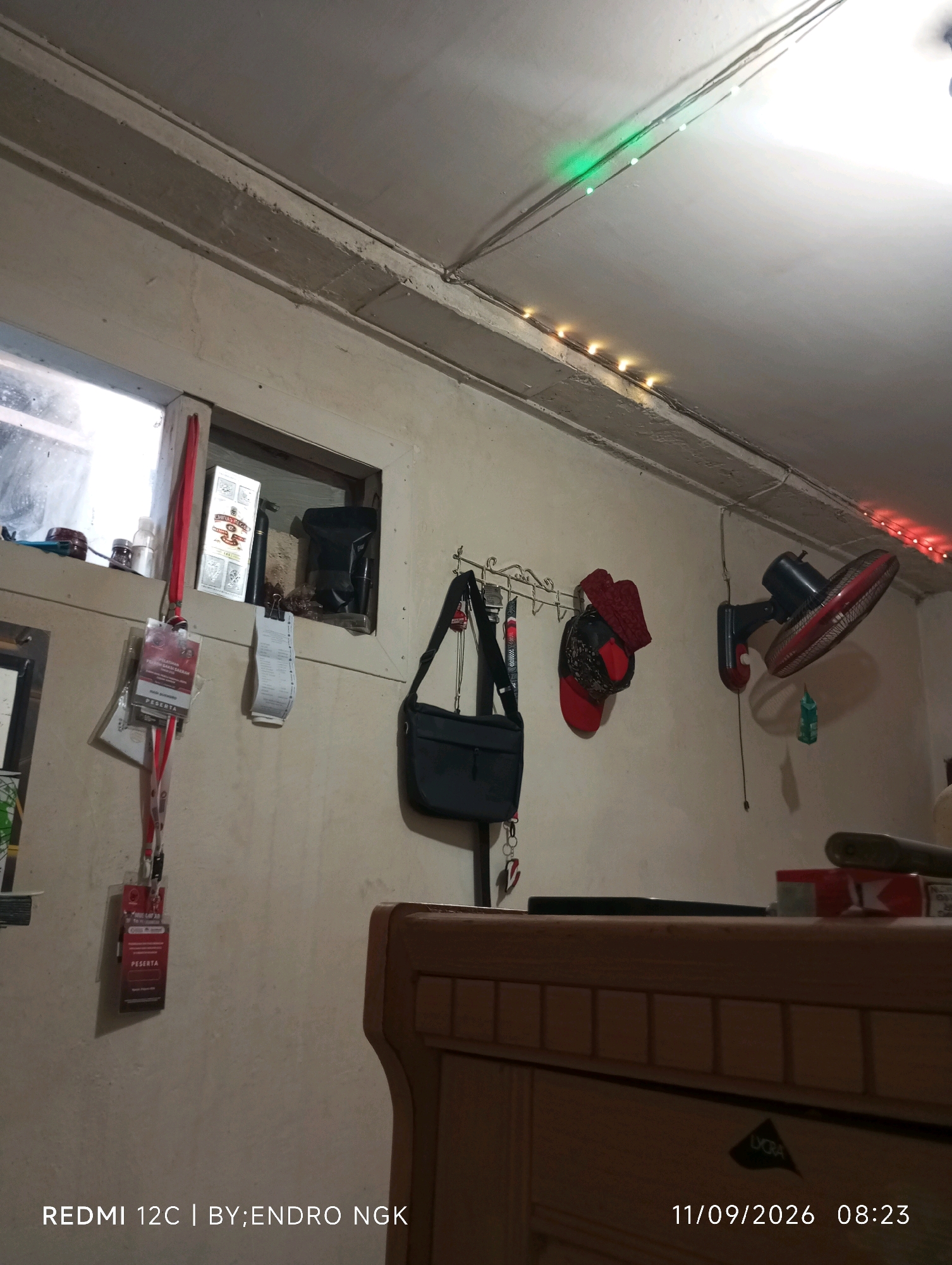 The image depicts an indoor scene of a room, likely a domestic or storage space. The walls are a neutral, off-white color and appear to be constructed from concrete or plaster. A visible seam runs horizontally across the upper portion of the wall, suggesting a join between building materials or a structural element.
On the left side of the image, there is a window opening with a view into an adjacent, darker space. This area contains various objects, including what appears to be a red tin, a bottle with a white cap, and a black bag or sack. A white vertical object, possibly a receipt or a tag, is hanging from a string attached to the window frame.
Below the window, a set of identification cards or lanyards are hanging, one of which is red and prominently displays the word "PESERTA" in white lettering.
On the right side of the wall, a black electric fan with red accents is mounted. It is positioned above a wall hook from which two red hats or caps are hanging. A black handbag is also suspended from a hook on the wall between the window and the fan.
The ceiling is also visible and appears to be made of similar material to the walls, with visible conduits and wiring. There are strings of small LED lights attached to the ceiling, with some lights emitting green and others a warm yellow or orange hue. Further to the right, a string of red LED lights is also visible.
In the foreground, a wooden piece of furniture, possibly a cabinet or desk, is partially visible. It is made of light-colored wood with a visible grain and a decorative pattern of rectangular inlays. A dark, flat object, perhaps a tablet or book, rests on the surface of this furniture. Towards the right edge of the furniture, a red and white package, possibly of cigarettes, is present. A green object, possibly a small bottle or pouch, hangs from a string near the fan.
The bottom of the image contains text overlay indicating the device used for capture "REDMI 12C | BY: ENDRO NGK" and the date and time "11/09/2026 08:23". The overall impression is of a functional, possibly cluttered, indoor environment.