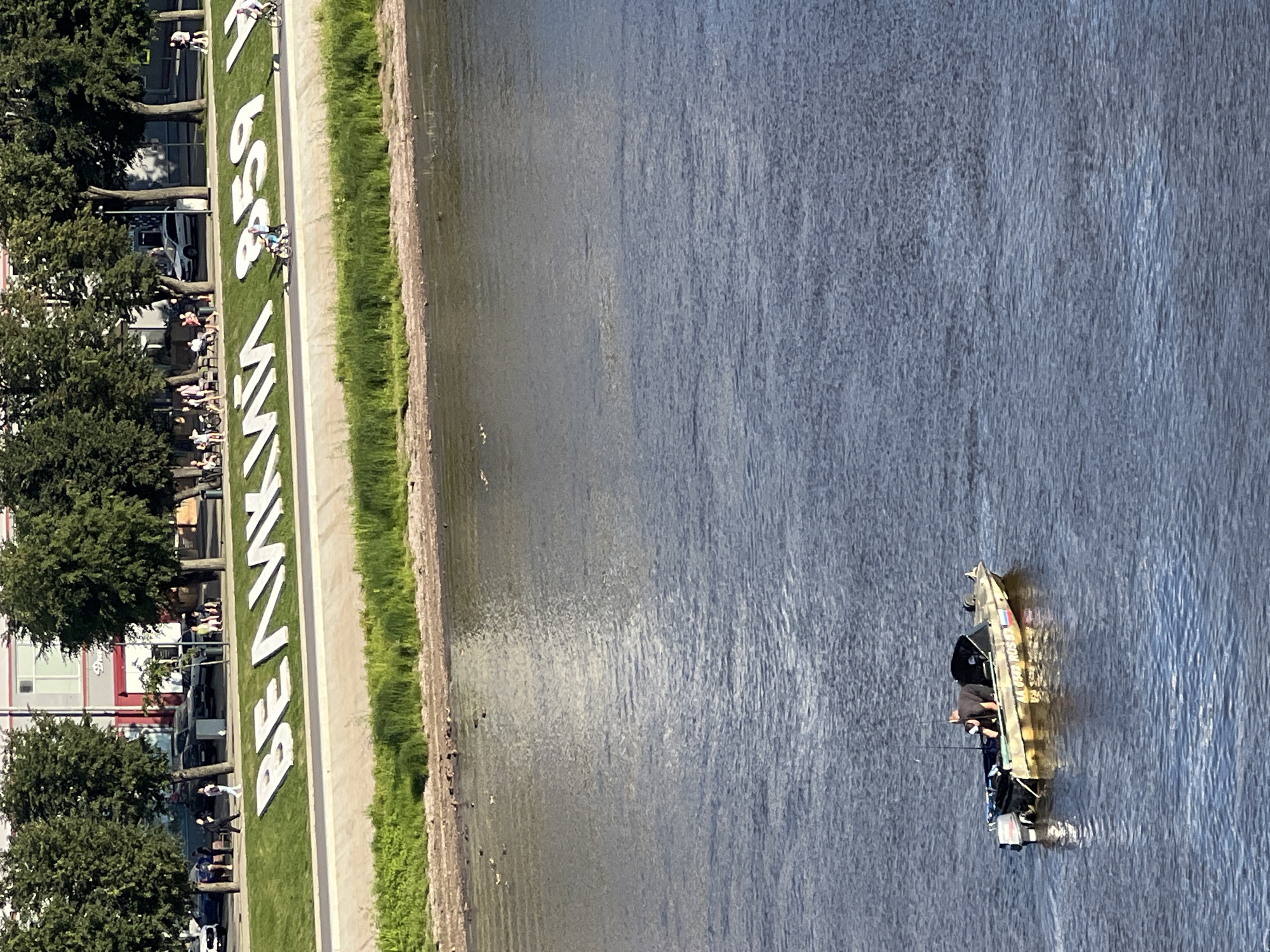 The image depicts a wide river flowing through the city of Veliky Novgorod, Russia. On the left bank, a pedestrian walkway lined with trees separates the river from a grassy embankment. Large white letters spelling out "ВЕЛИКИЙ НОВГОРОД" are prominently displayed on the embankment, alongside the numbers "859". Several people are visible walking and cycling along the walkway. In the river, a small motorboat with two people aboard is navigating the water. Ducks are also scattered across the river's surface. The water is calm with ripples reflecting the sunlight. The overall scene suggests a pleasant day in the city, with both recreational activities and historical landmarks visible.
