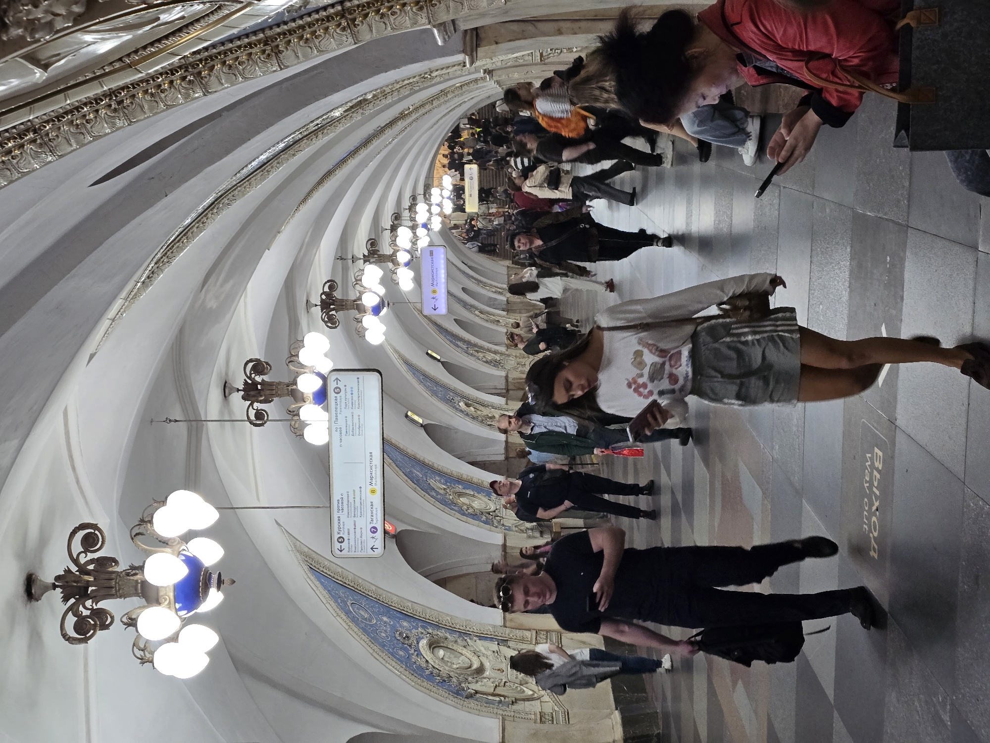 Here is a detailed description of the image:
**Location:** The image captures the interior of a metro station, possibly in Moscow, Russia. The architecture is grand and ornate, suggesting a historical or centrally located station.
**Environment:** The station features a curved ceiling with white panels and gold detailing. Elaborate chandeliers with blue accents hang along the corridor. Blue and white decorative panels line the walls. The floor is gray and appears to be tiled, with a "Way out" or "Выход" sign in Russian visible.
**People:** The image shows several people walking through the station. They are diverse in age and attire, suggesting a mix of commuters and visitors. Many are looking at their phones.
**Actions and Interactions:** People are walking and moving through the station. Some seem to be navigating using their phones. The general impression is of a busy transportation hub with people going about their daily routines.
**Overall Impression:** The scene conveys the impression of a grand and busy metro station, with a blend of historical architecture and modern commuters.