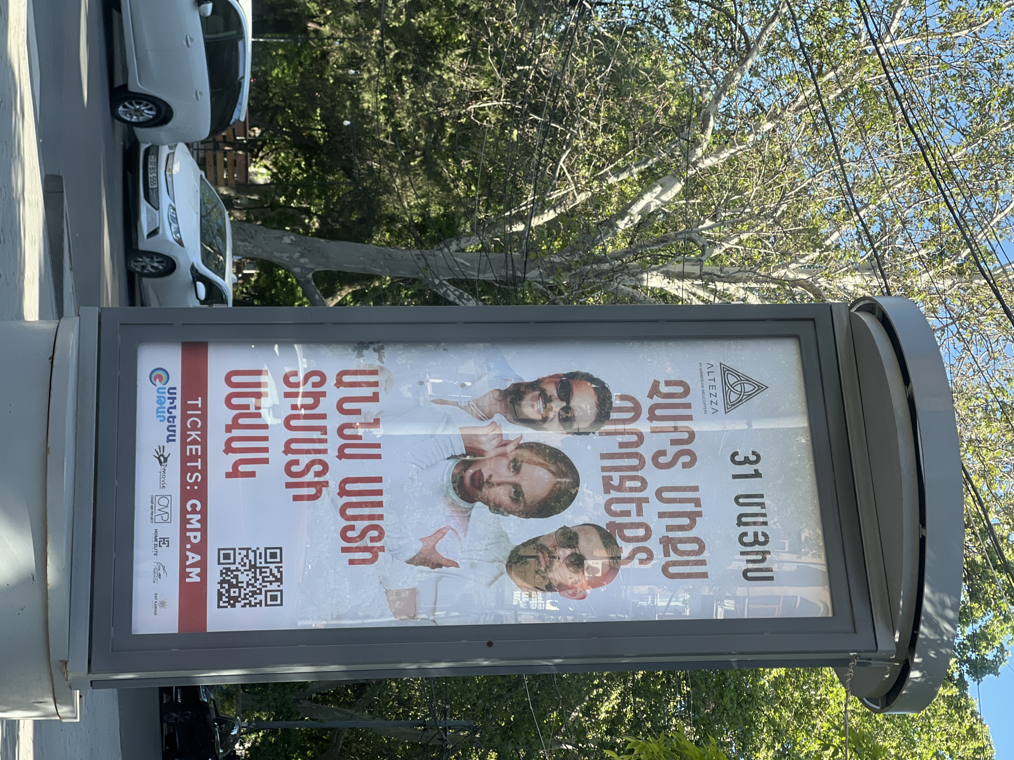 The image depicts a gray rectangular bus stop advertising poster in Yerevan, Armenia. The poster features the date "31 ՄԱՅԻՍ" (May 31st), the word "ALTEZZA," and "BY ARMENIAN HELICOPTERS." It promotes "ՉԱՐՏ ՄԻՔՍ ՊՐՈՋԵՔՏ" (Chart Mix Project), followed by "ԱՆՆԱ ԱՍՏ" (Anna Ast) and the names "ՏԻՄԱՏԻ ՍԵՎԱԿ" (Timati Sevak). A QR code and website "TICKETS: CMP.AM" are displayed. The poster includes logos of various entities, "ՍԻՆԵՄԱ ՍՏԱՐ," "Curur," "HOME ELITE PROJECT," and "RAF KANIAN." Behind the bus stop, there are trees and a clear blue sky. Parked cars are also visible.