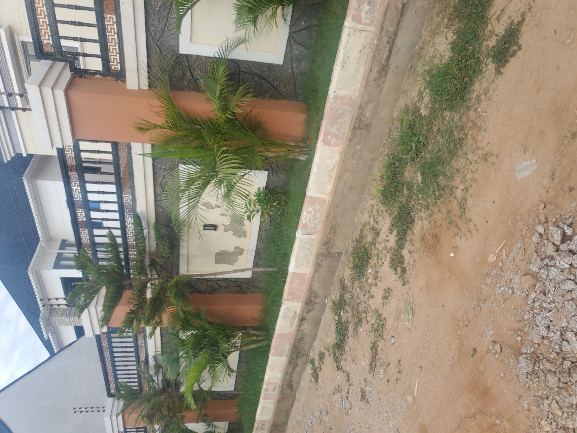The image shows a residential property with a fence and a house in Barden Yana, Nigeria. The fence is made of concrete pillars with a black metal railing. Behind the fence, there are palm trees and a house with a dark roof and white walls. The area in front of the fence is a dirt road with some gravel and scattered vegetation. A drainage ditch runs along the edge of the property. The sky is overcast.