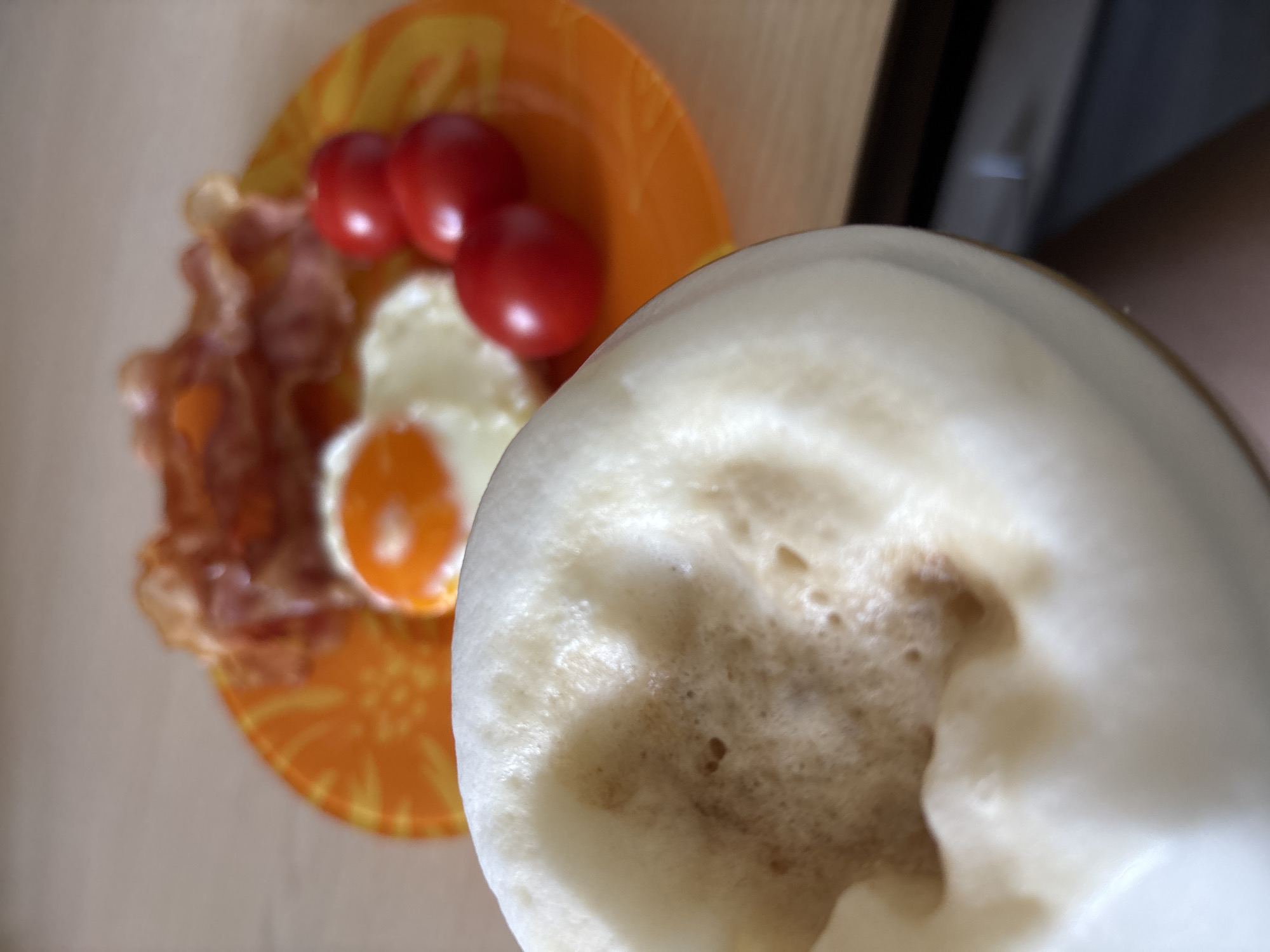 A close-up shot presents a meal and a drink on a wooden surface. An orange plate holds bacon strips, a fried egg, and three cherry tomatoes. A drink with a thick layer of foam fills the right side of the frame, obscuring part of the plate. The background appears blurred, suggesting a domestic setting. No clear indication of Rusanovo, Russia, is apparent in the image.