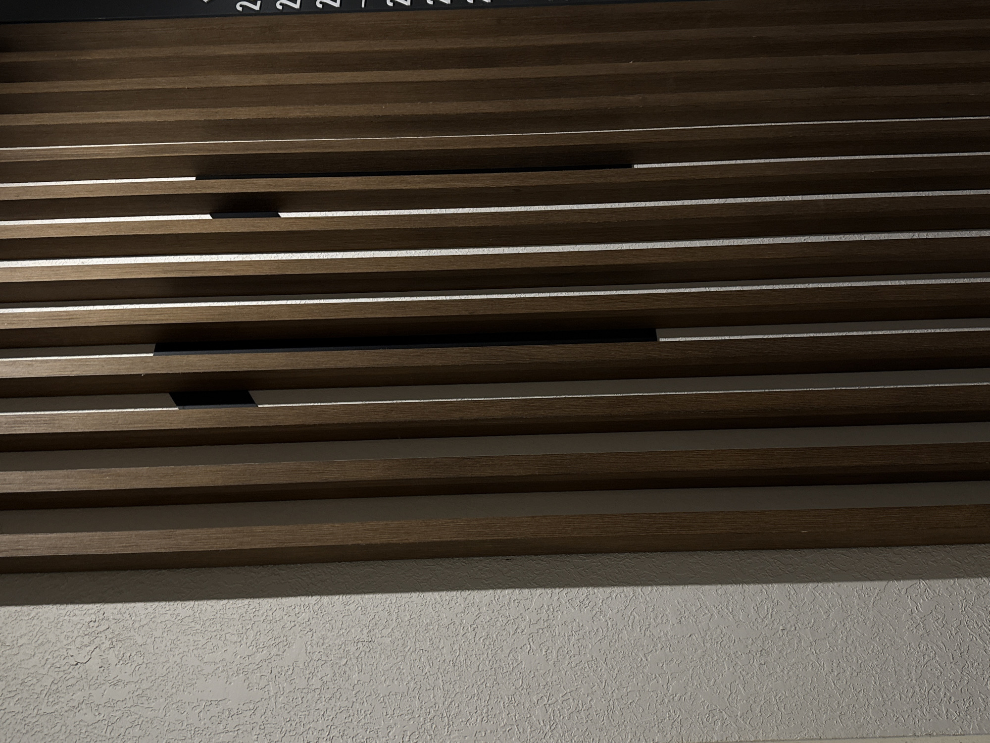A textured white wall is juxtaposed with a vertical wooden slat wall. The wooden slats are dark brown and have a subtle sheen, with thin gaps between them revealing a white surface behind. A black object is attached to the wooden slat wall, and it is partially visible between two slats. On the far right edge of the image, a black surface with white lettering is visible. The lettering appears to be a list of numbers, such as "2", "2", and "2", arranged vertically. The lighting suggests an indoor environment, possibly a hallway or a modern interior space. The scene is static, with no people or apparent activity.