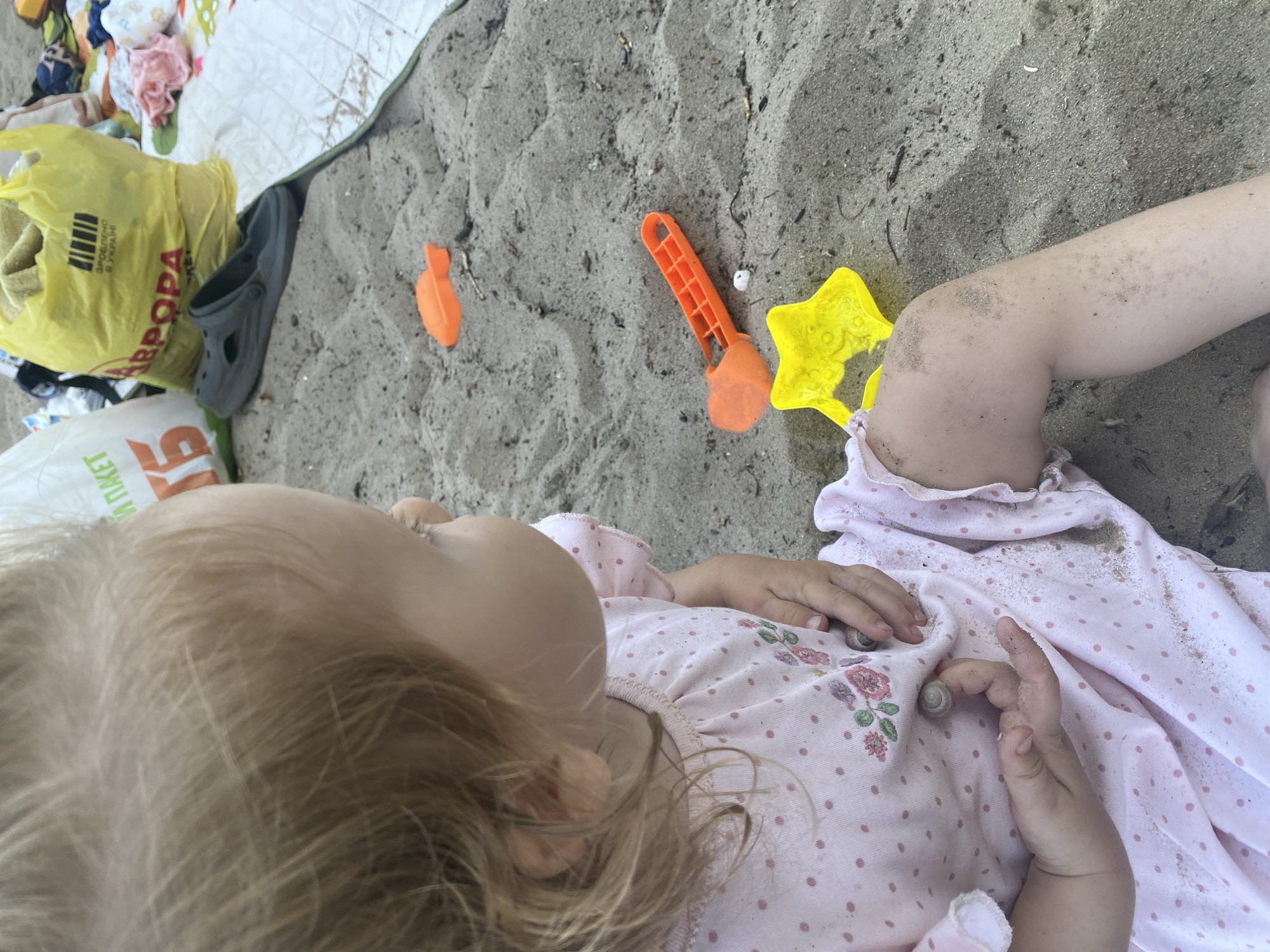 A young child with blonde hair, wearing a pink dress with polka dots and floral embroidery, sits on a sandy beach. The child's hands, covered in sand, hold small seashells. Several brightly colored children's beach toys, including an orange fish-shaped shovel and a yellow star-shaped mold, are scattered in the sand around the child. In the background, a yellow plastic bag and a grey sandal are visible, suggesting a recreational setting. The overall scene conveys a typical day at a beach.
