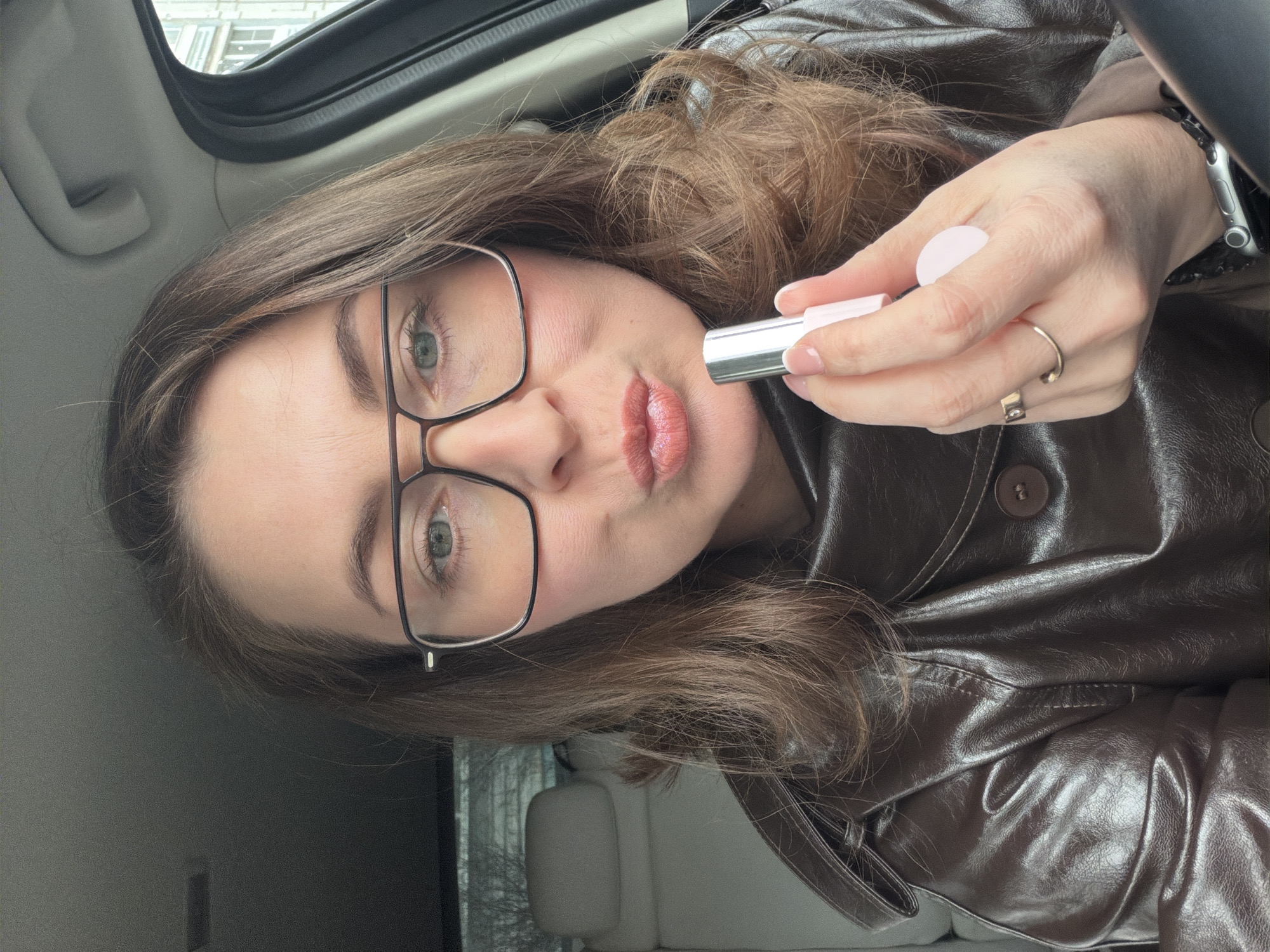 A fair-skinned woman with light brown hair is sitting inside a car and applying lipstick. She is wearing glasses with black frames and a brown leather jacket. Her lips are puckered as she holds a small, silver lipstick tube with a white cap in her right hand. The car's interior is visible in the background, showing a light-colored seat and parts of the vehicle's structure. The location is identified as Unknown, Unknown.