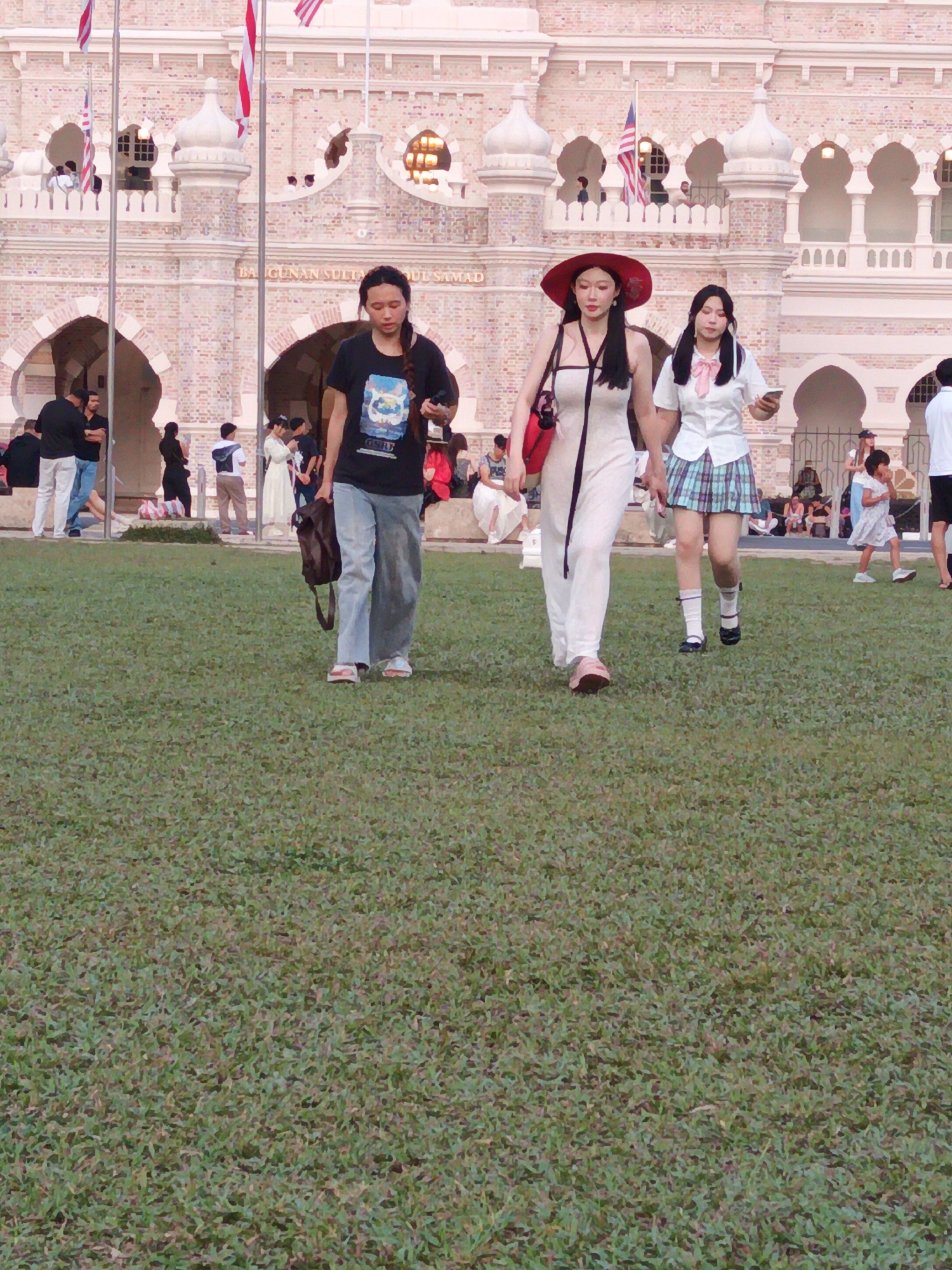 The image shows a group of people in front of the Sultan Abdul Samad Building in Kuala Lumpur, Malaysia. The building is a prominent landmark in the city, known for its colonial architecture and historical significance. In the foreground, three young women are walking on a grassy lawn. The woman on the left is wearing a black t-shirt and jeans, carrying a brown bag. The woman in the middle is wearing a white dress and a red hat, with a red bag slung over her shoulder. The woman on the right is wearing a school uniform, consisting of a white shirt, a plaid skirt, and black shoes. Several other people are visible in the background, some walking, some standing, and some sitting. The scene appears to be a public gathering or tourist attraction, with people enjoying the outdoor space and the historical architecture. The lighting suggests it is daytime.