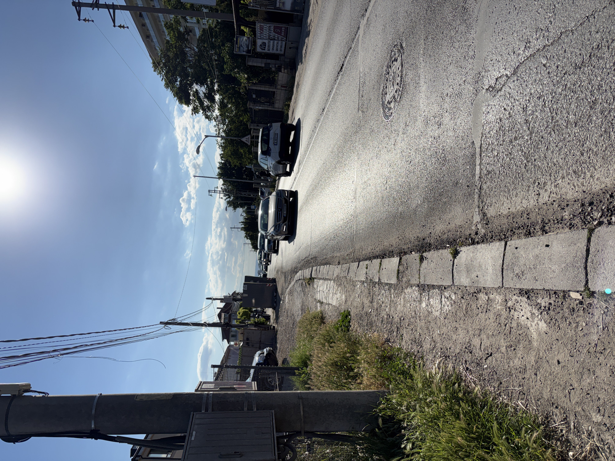 The image depicts a street scene in Varna, Bulgaria, with a focus on the roadside and the road itself. The perspective is from a sidewalk or unpaved area adjacent to the road.
In the foreground, to the left, is a concrete utility pole with various wires and cables attached to it. A metal utility box is mounted on the pole. Beside the pole, there is a patch of dry grass and some weeds growing. The ground consists of dirt and gravel.
The road, paved with asphalt, curves to the right and recedes into the distance. Several cars are driving on the road. The sunlight is strong, casting reflections on the road surface and creating lens flare in the sky. The sky is blue with some scattered white clouds.
On the right side of the road, there are fences, trees, and buildings. One building appears to be a multi-story residential or commercial structure with balconies. Another area on the right features advertisements or signs.
In the mid-ground, more cars are visible on the road. A manhole cover is present on the asphalt. The sidewalk or paved edge of the road is made of concrete slabs.
The overall impression is of a sunny day on an urban or suburban street, possibly in a less developed or older part of the city, given the presence of overhead wires and the condition of the road and sidewalk. The area appears to be a transitional zone between a residential or commercial area and a more open space.