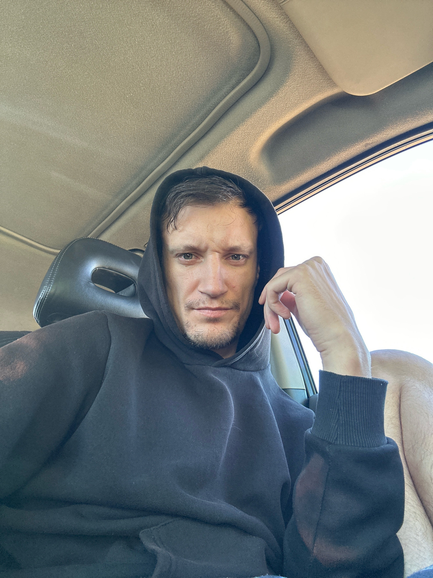 A man is sitting in the driver's seat of a car, wearing a black hoodie and facing the camera. His right hand is resting on his chin, and he has a serious expression on his face. The interior of the car is visible, including the headrest of the seat next to him and the ceiling of the car. The lighting suggests it is daytime, with sunlight coming in from the right side of the frame. The exact location within Makiv, Ukraine, is not discernible from the image.
