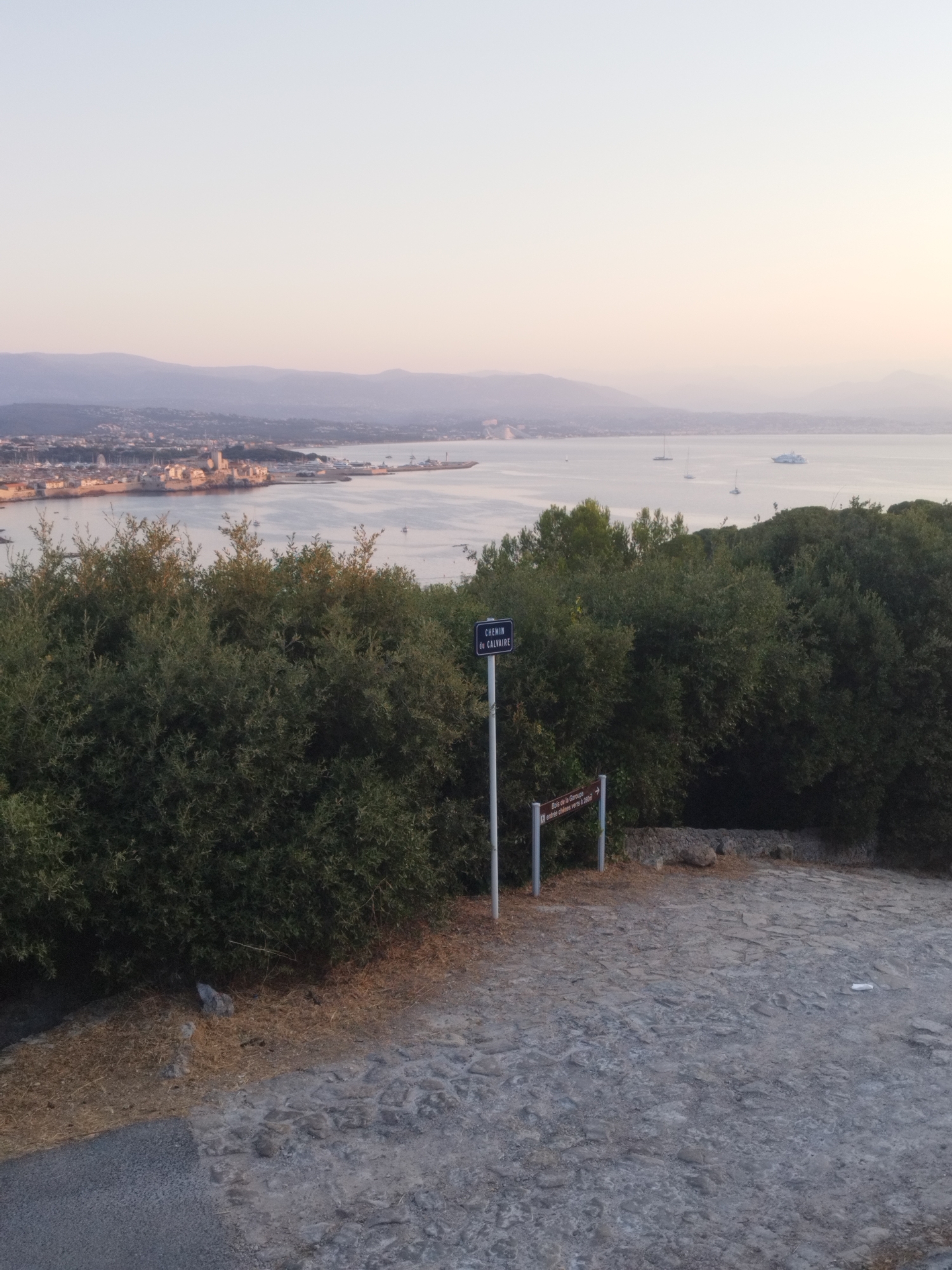 The image depicts a scenic view overlooking the bay of Antibes, France. In the foreground, a cobblestone path leads towards a dense green shrubbery. A signpost stands prominently, with one sign reading "CHEMIN du CALVAIRE" in blue, indicating a path. Another brown sign below it indicates directions to "Bois de la Garoupe" and "entre chênes verts à 200m".
Beyond the foliage, a panoramic vista of Antibes unfolds. The city, with its buildings and a prominent harbor filled with boats, stretches along the coastline. The calm sea, reflecting the soft hues of the sky, is dotted with several sailboats and a larger white vessel, possibly a cruise ship or a yacht. In the distance, hazy mountains form a backdrop against the pale, serene sky, which exhibits a gradient of light colors, suggesting either early morning or late afternoon. The overall atmosphere is tranquil and picturesque.