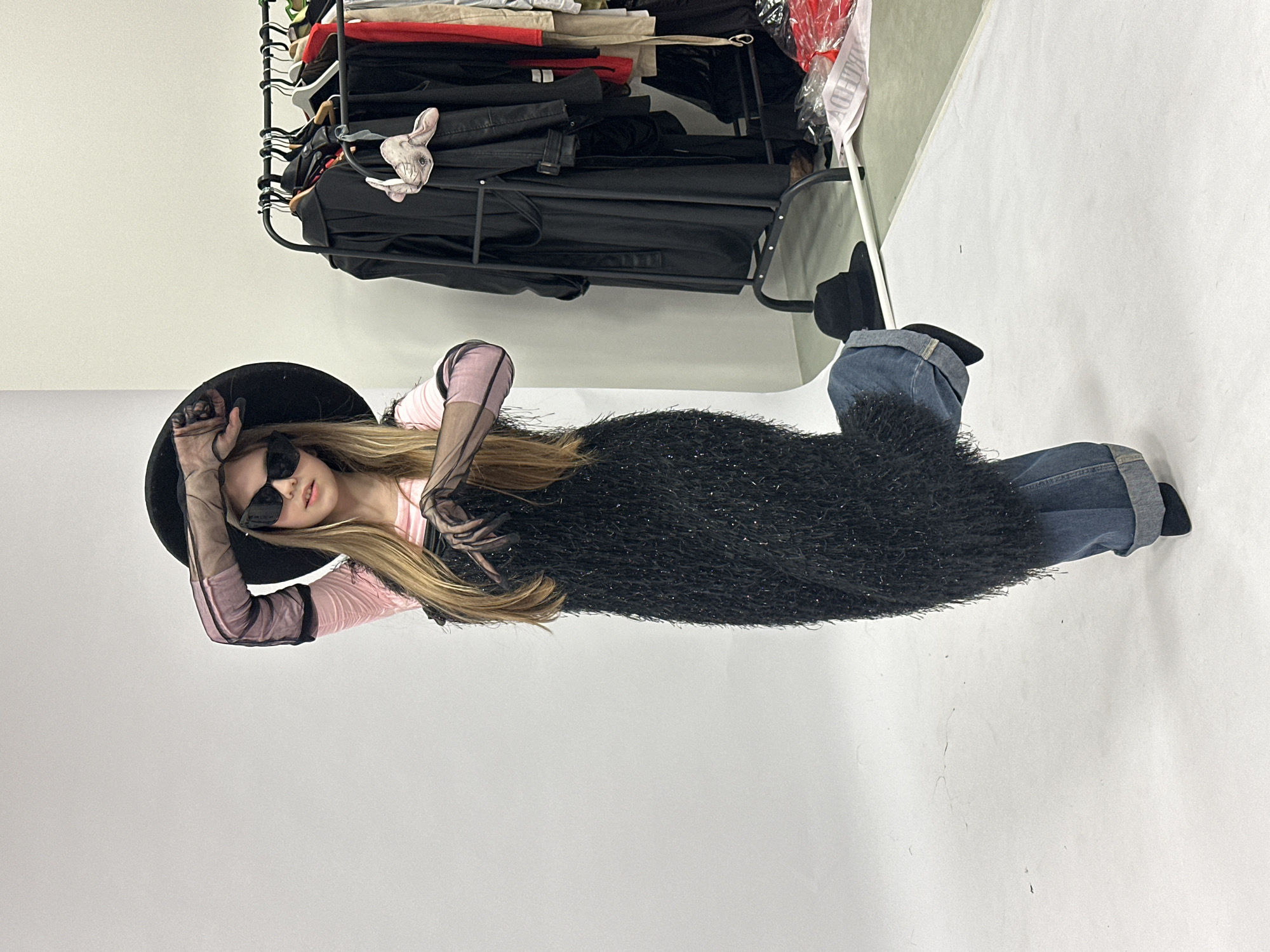 A woman is lying on a white surface, likely a photography studio floor, with a black fuzzy dress and blue jeans. She is also wearing a black hat, black sunglasses, and a pink top with sheer black sleeves. A clothing rack with various garments, including black leather jackets, is in the background. The location cannot be determined as Votkinsk, Russia, solely from the image.