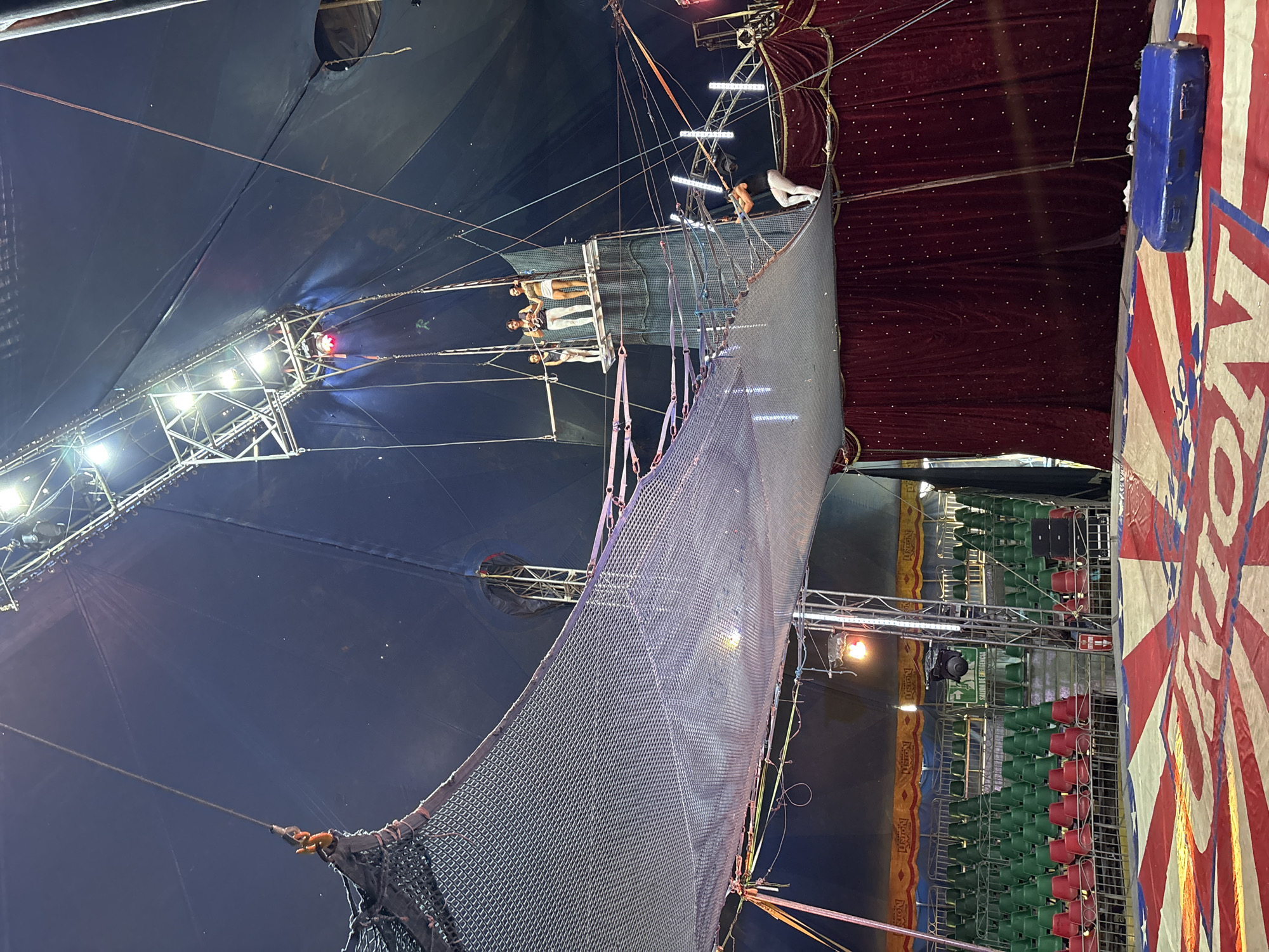 The image shows the interior of a circus tent in Tejalpa, Mexico. Several performers are positioned on a high trapeze platform. Below them, a large safety net is stretched out. Seating is visible to the right, some of which is red and some green. The tent is supported by a framework of metal poles and cables, with bright lights attached to the structure. The tent's roof is dark blue. A section of the tent is decorated with red and white stripes.