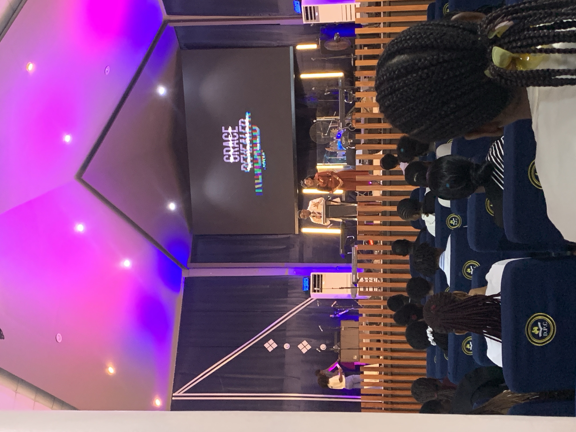 An indoor space, likely in Dar es Salaam, Tanzania, features rows of people seated in blue upholstered chairs with yellow emblems, potentially attending a presentation or service. A screen displays "Grace Revealed," and two people are on stage. The room has a geometrically shaped ceiling with pink and blue lighting, and wooden slats line the walls. There are cooling units and musical instruments visible.