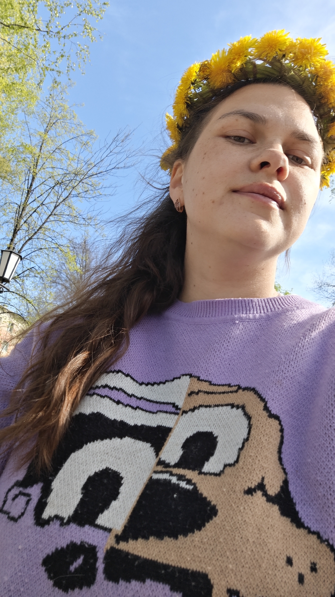 A young woman poses for a selfie outdoors. She wears a crown of yellow dandelions and a lilac sweater with a cartoon dog design. Her dark hair falls to one side. A lamppost is visible in the background along with green trees and a blue sky. The photo was taken in Chaikovsky, Russia.