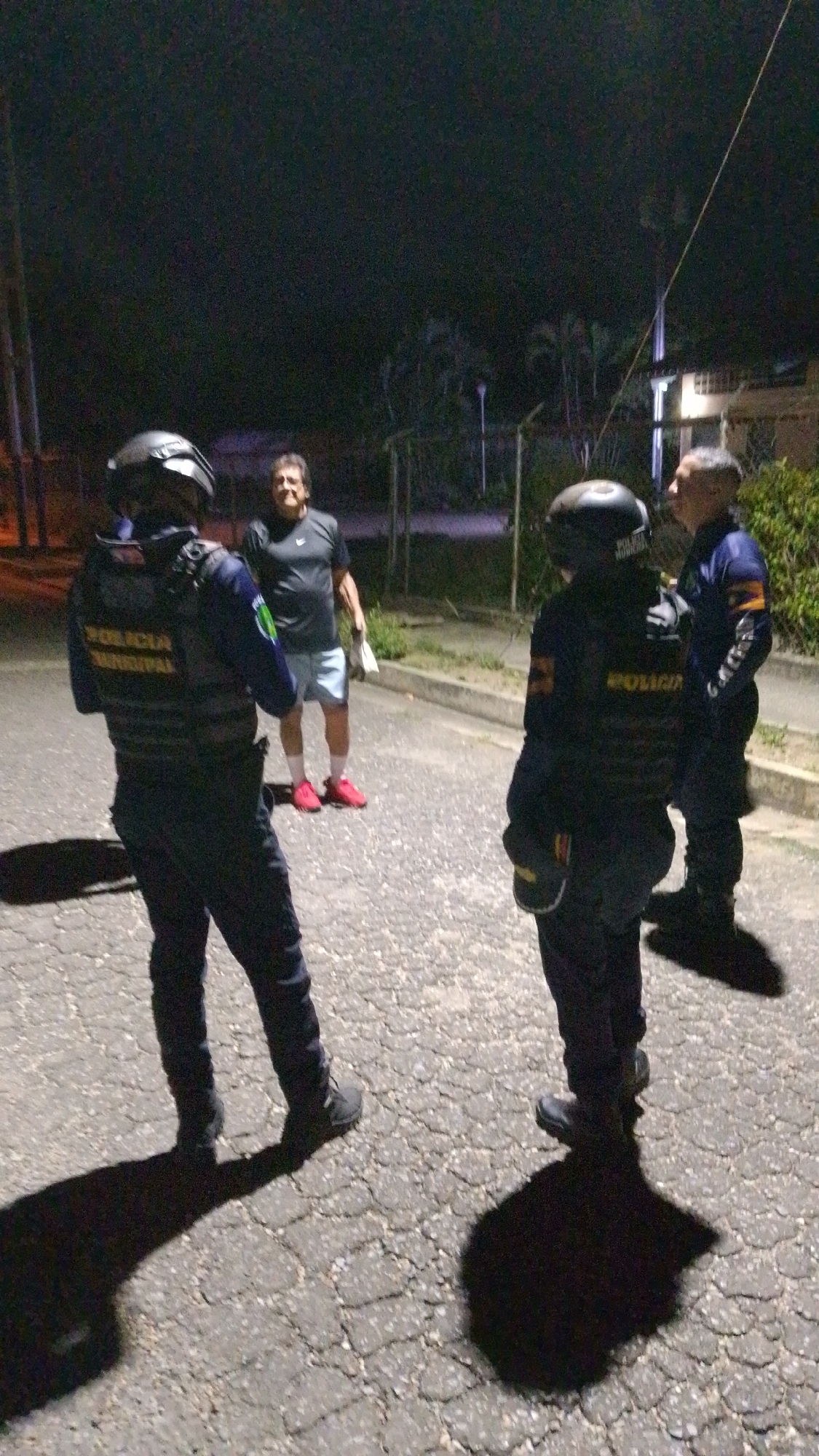 The image shows three uniformed police officers and one civilian male standing on a paved road, possibly in Tinaquillo, Venezuela. The two officers closest to the camera wear dark protective gear including helmets and vests with "POLICIA MUNICIPAL" visible on the back of one. The third officer stands to the right. The civilian male wears a dark t-shirt, light-colored shorts, and red shoes. He is holding something white in his left hand. The scene is dimly lit. Background vegetation and a building are visible.