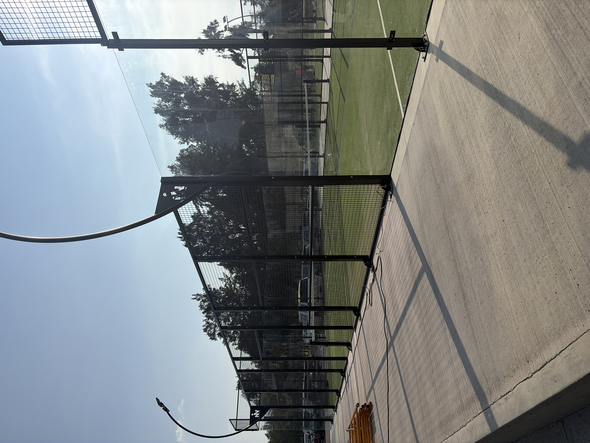 The image shows a partial view of a paddle tennis court in Puebla, Mexico. The court is enclosed by a wire mesh fence and glass panels. The court surface is green with white lines. Outside the court, there's a concrete walkway. The sky is visible in the background along with trees and parked cars. In the foreground, there's a yellow object, possibly a rolled-up net or tarp, and a light pole. No people are clearly visible in the image. The perspective is from a low angle, looking upwards.