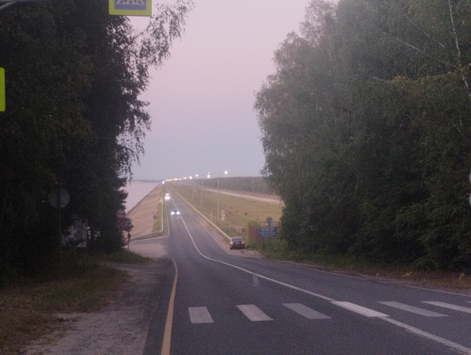 The image depicts a dimly lit road, likely at dusk, bordered by dense trees on both sides. A paved road with white lane markings and a pedestrian crossing is visible. Several vehicles are present: two cars are driving on the road in the distance, and another car is parked on the right shoulder. Streetlights line the road in the background, illuminating the scene with a soft glow. A body of water, possibly a lake or river, is visible on the left side of the road, separated by a grassy embankment and trees. A pedestrian crossing sign is visible on the left. The overall atmosphere is calm and serene, with a touch of mystery due to the low light conditions. The location is identified as Urkovo, Russia.