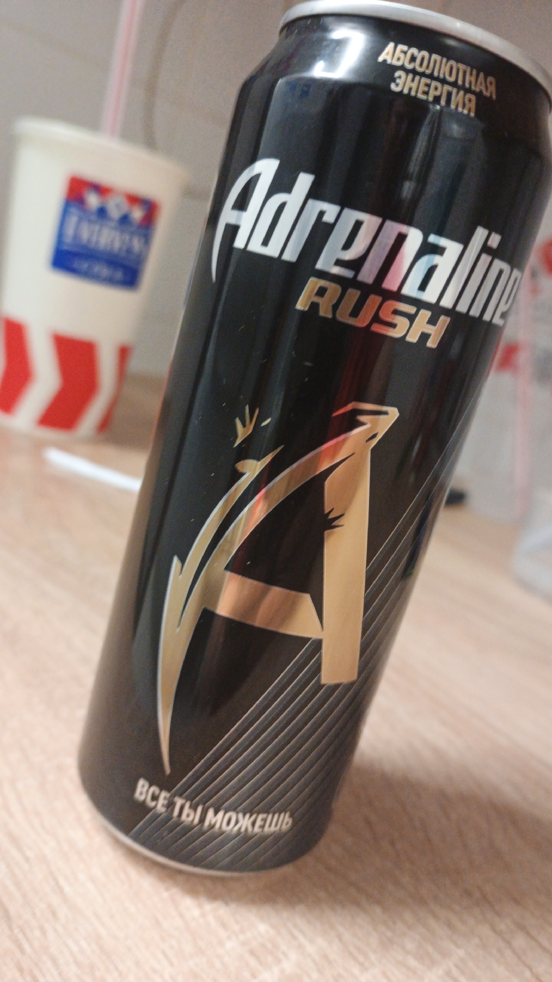 The image is a close-up, slightly angled shot of a black can of Adrenaline Rush energy drink. The can is positioned on a light-colored wooden surface. The text on the can is in Russian, with "Adrenaline Rush" prominently displayed in silver lettering. Above this, in smaller silver text, is "АБСОЛЮТНАЯ ЭНЕРГИЯ" (Absolute Energy). Near the bottom of the can, in white text, is "ВСЕ ТЫ МОЖЕШЬ" (You can do anything). A stylized gold logo resembling a soaring bird or an abstract 'A' with wings is also visible on the can. In the blurred background to the left, a white disposable cup with red chevron stripes and a blue logo containing red and white elements is visible, along with a pink straw. The scene appears to be an indoor setting, possibly a kitchen or a desk area, given the wooden surface and the presence of a disposable cup. There are no people or animals in the image. There is no information that allows to place this image in the city of Pavlovsky Posad, Russia.