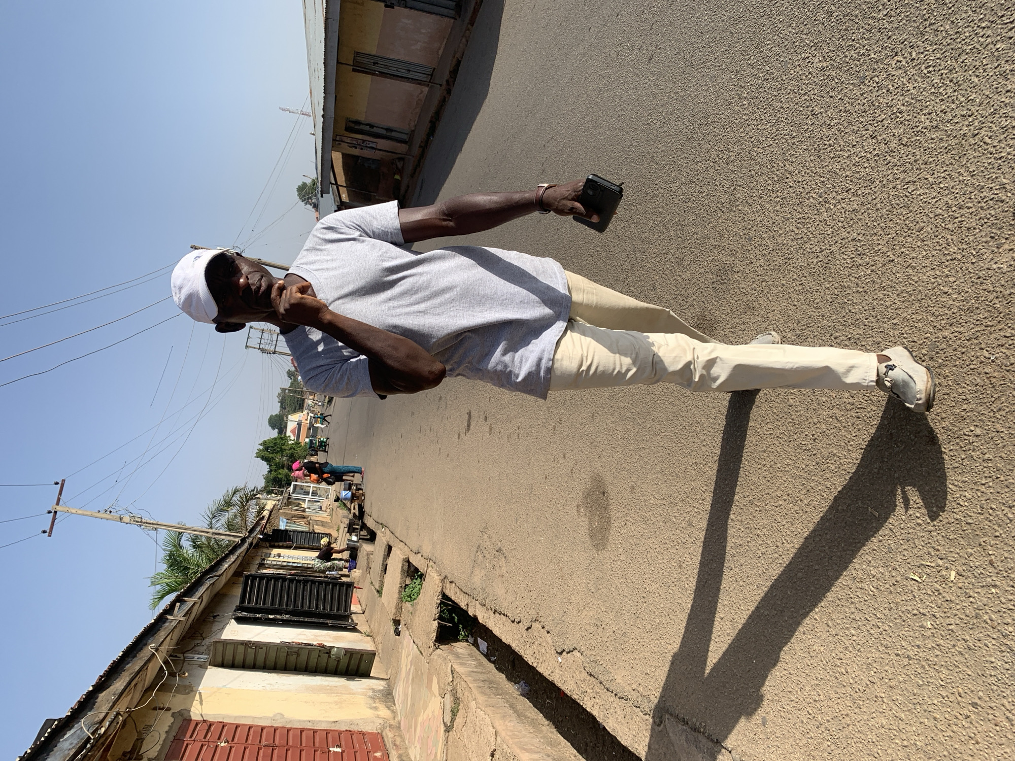 An adult male stands in the middle of a paved street in Jos, Nigeria. He wears a light gray t-shirt, tan pants, and white shoes, and a white cap. He is holding a black smartphone in his right hand and touching his face with his left. The street is flanked by low buildings, some with corrugated metal roofing and closed shutters, and at least one with a red exterior. There are other people further down the street to the left, possibly vendors or customers in front of shops. Power lines are visible overhead. The sky is bright and there is strong sunlight, casting long shadows on the street. A drainage ditch runs along the side of the street.