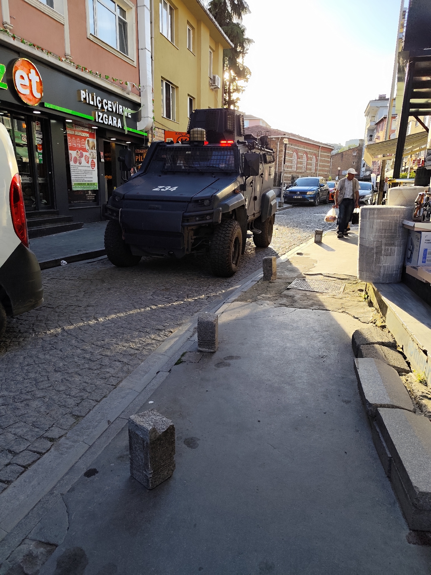 Here's a forensic description of the image:
**Setting:** Outdoor urban environment, Sarıyer, Turkey. The scene is a cobblestone street with buildings on either side.
**Objects:**
* An armored vehicle with the number "234" prominently displayed on the front.
* A white van partially visible on the left.
* Several parked cars further down the street.
* Stone bollards lining the edge of the sidewalk.
* A store with a sign that reads "PİLİÇ ÇEVİRME IZGARA".
* A partially-covered stall on the right.
**People:**
* An elderly man is walking down the street, carrying bags. He is wearing a light-colored hat and jacket.
**Environment:**
* Cobblestone street is visible
* The weather appears to be sunny.
* There is a building with a light green exterior and red tile roof in the background.
* The sun is high in the sky, creating strong shadows.
