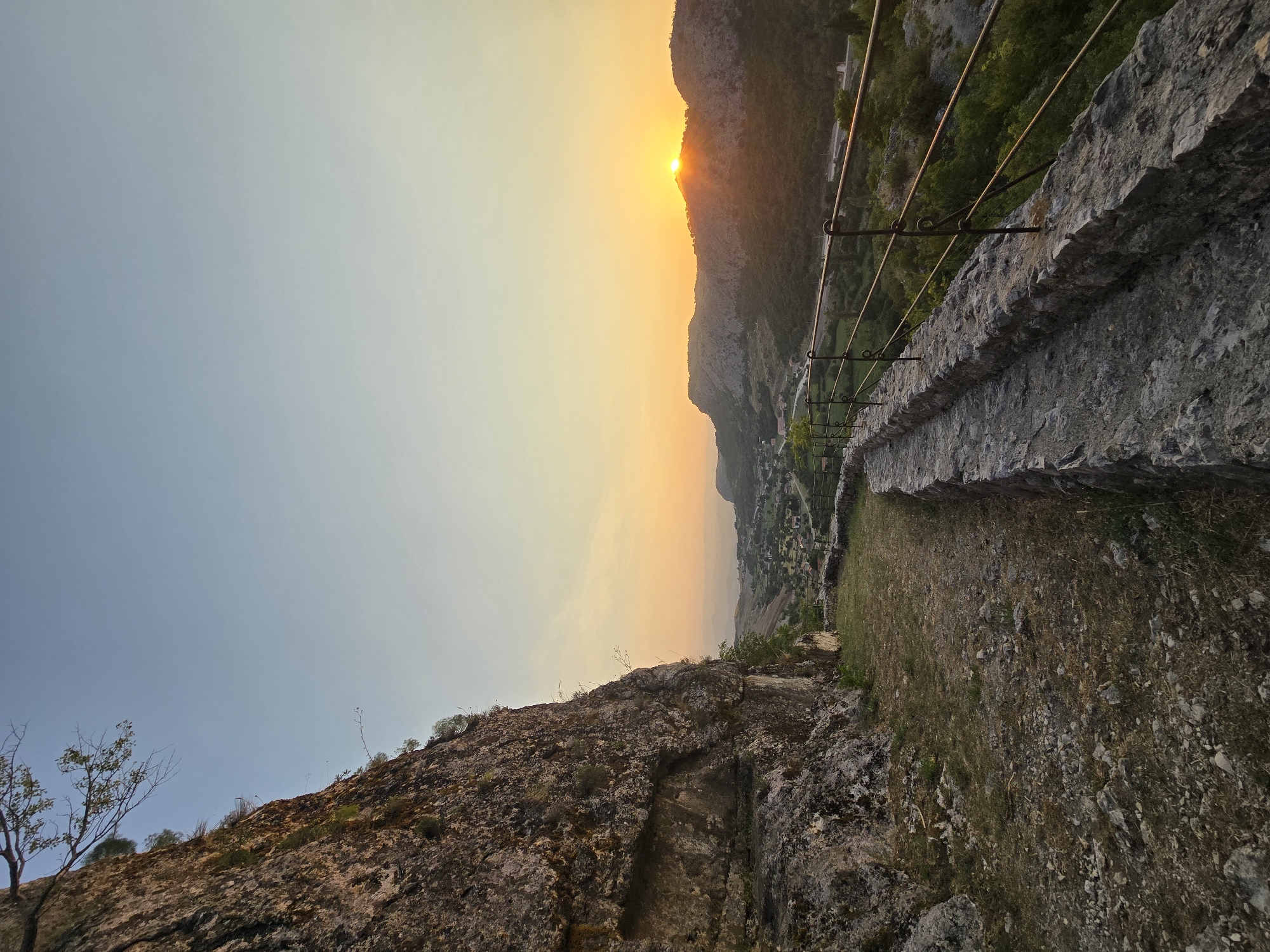 The image captures a sunset view from a rocky cliffside, overlooking a valley with a village and mountains in the distance. A stone pathway with a metal railing follows the edge of the cliff. The sky is clear with a gradient of blue and orange hues. The sun is setting behind the mountains, casting a warm glow on the landscape. The village appears to be nestled among rolling hills and lush vegetation. The pathway is made of irregular stones and is bordered by a low stone wall topped with a dark metal railing. The cliff face to the left is rugged and textured, with sparse vegetation clinging to its surface. The environment suggests an outdoor, elevated viewpoint, likely a historical or natural landmark.