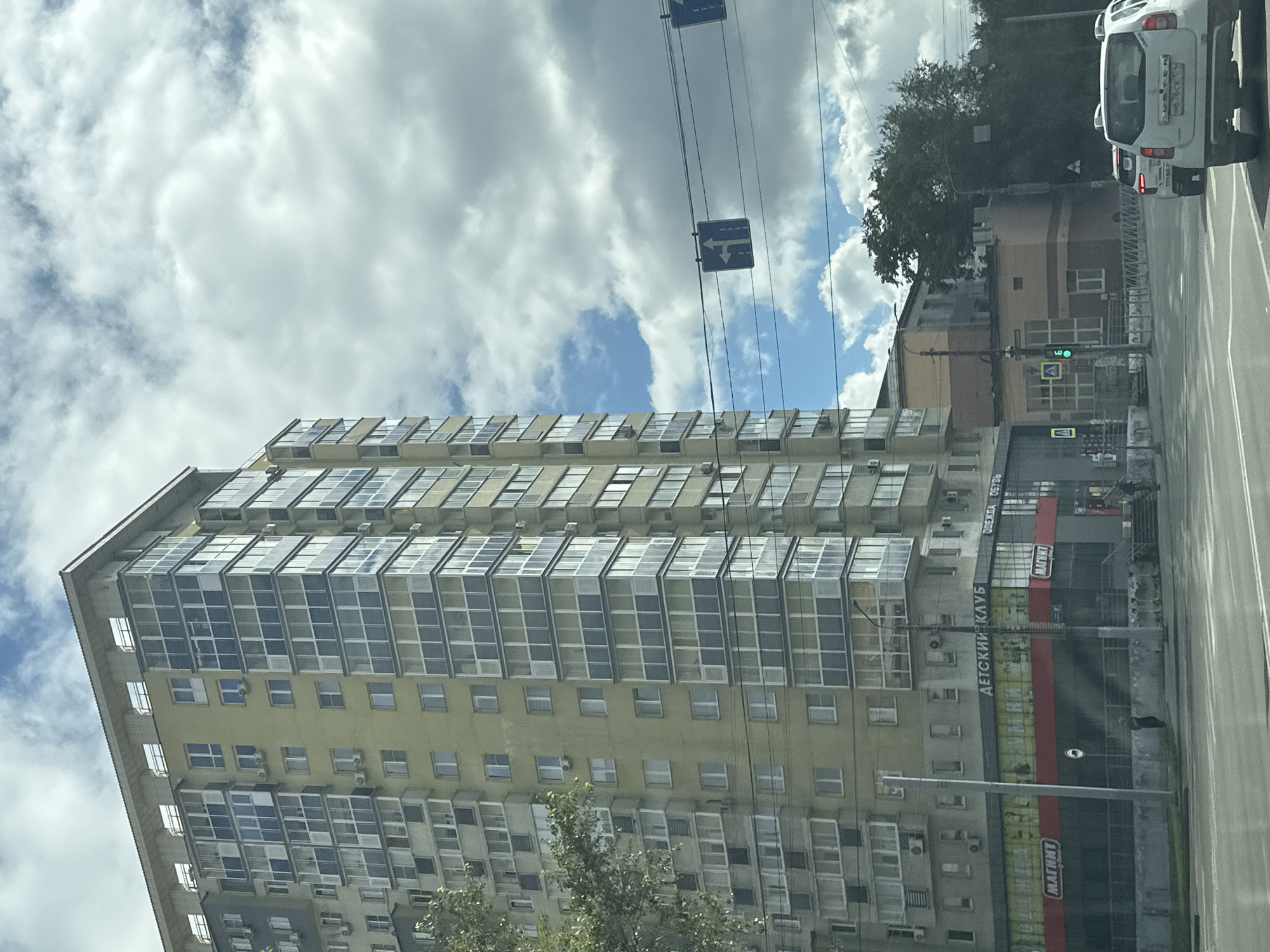 The image shows a tall, multi-story apartment building with balconies on the left side of the frame. The building is painted a light yellow and has many windows. In front of the building, there is a street with cars. There are also traffic signs visible, including a sign indicating left and straight ahead. A retail store with a red and black facade is at the base of the building, with signs reading "МАГНИТ" and "ДЕТСКИЙ КЛУБ". The sky is cloudy with patches of blue.