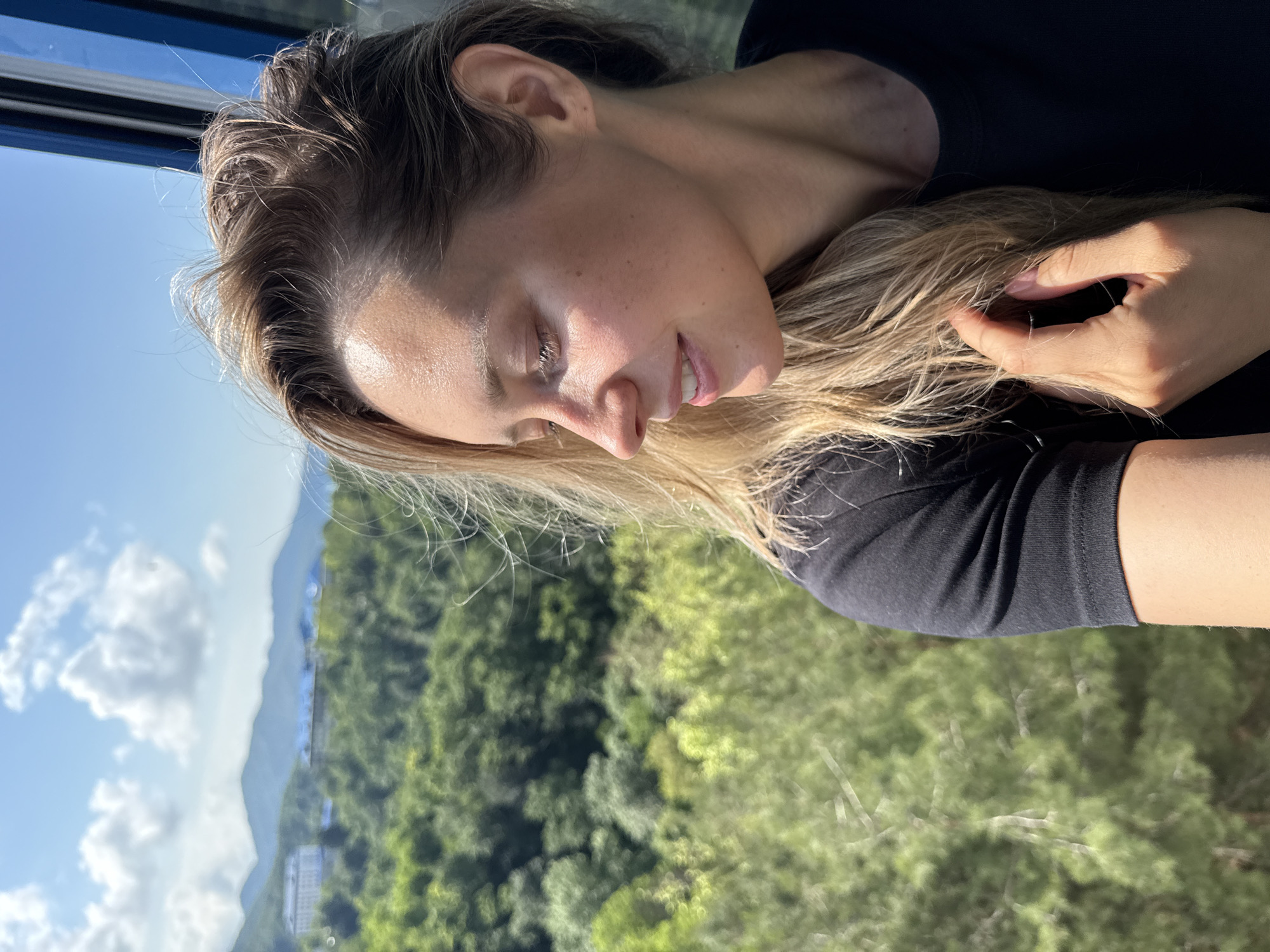 In the city of Nebug, Russia, a young woman with blonde hair is captured from the chest up, looking to her left. She is wearing a black t-shirt. Her right hand is gently touching her hair, which is illuminated by sunlight. The background is a lush green forest with distant mountains and a blue sky with white clouds, suggesting an outdoor setting, possibly from a cable car or high vantage point.