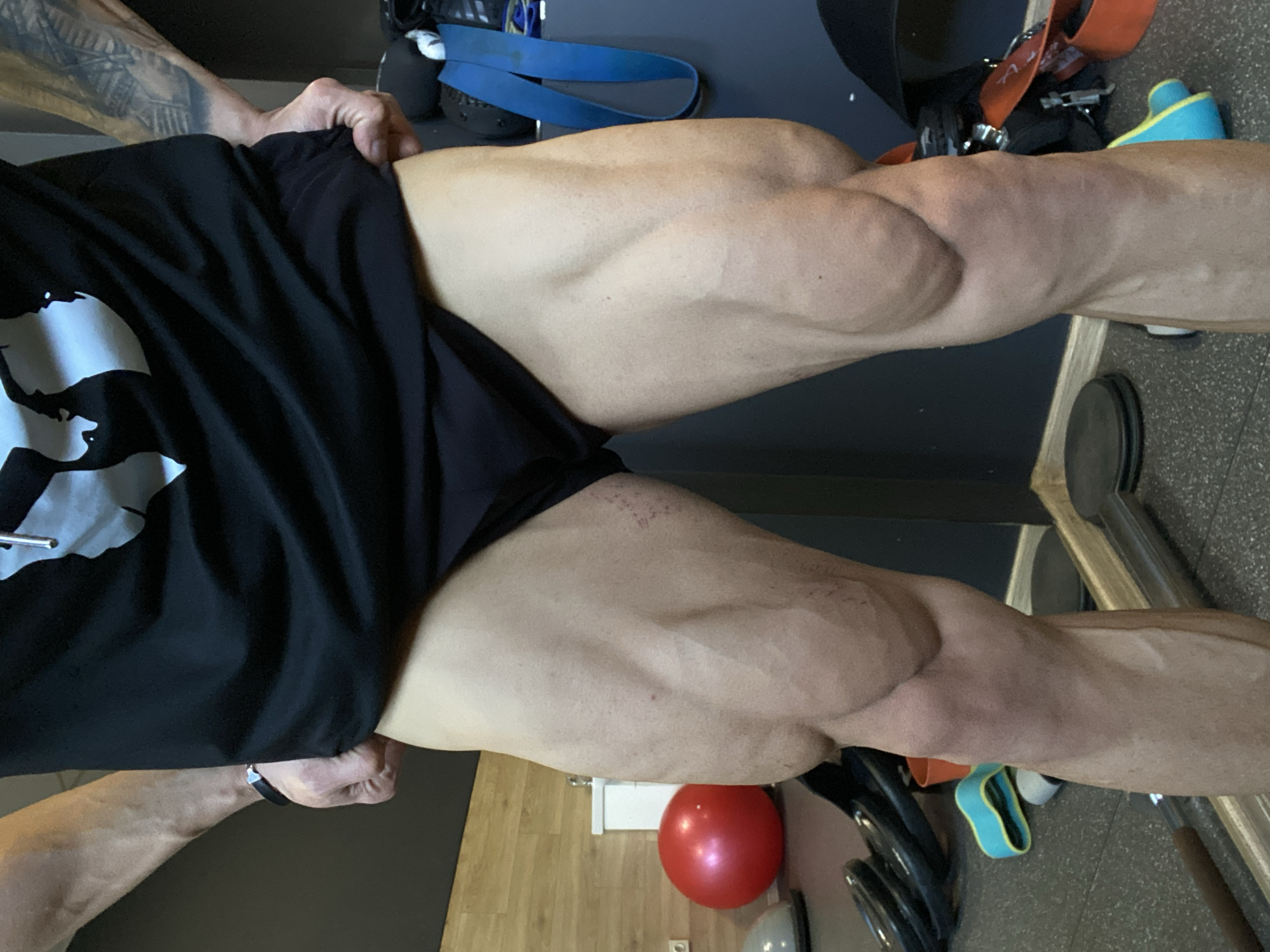 The image depicts a person's legs in what appears to be a gym environment. The person is wearing black shorts. The legs are muscular with visible veins. The background includes gym equipment such as weights and resistance bands. The flooring appears to be wood laminate and another section looks like grey gym flooring. There are also a red ball, a white electrical box and a power outlet. The location is not identifiable as Pleven, Bulgaria based on visual information alone.