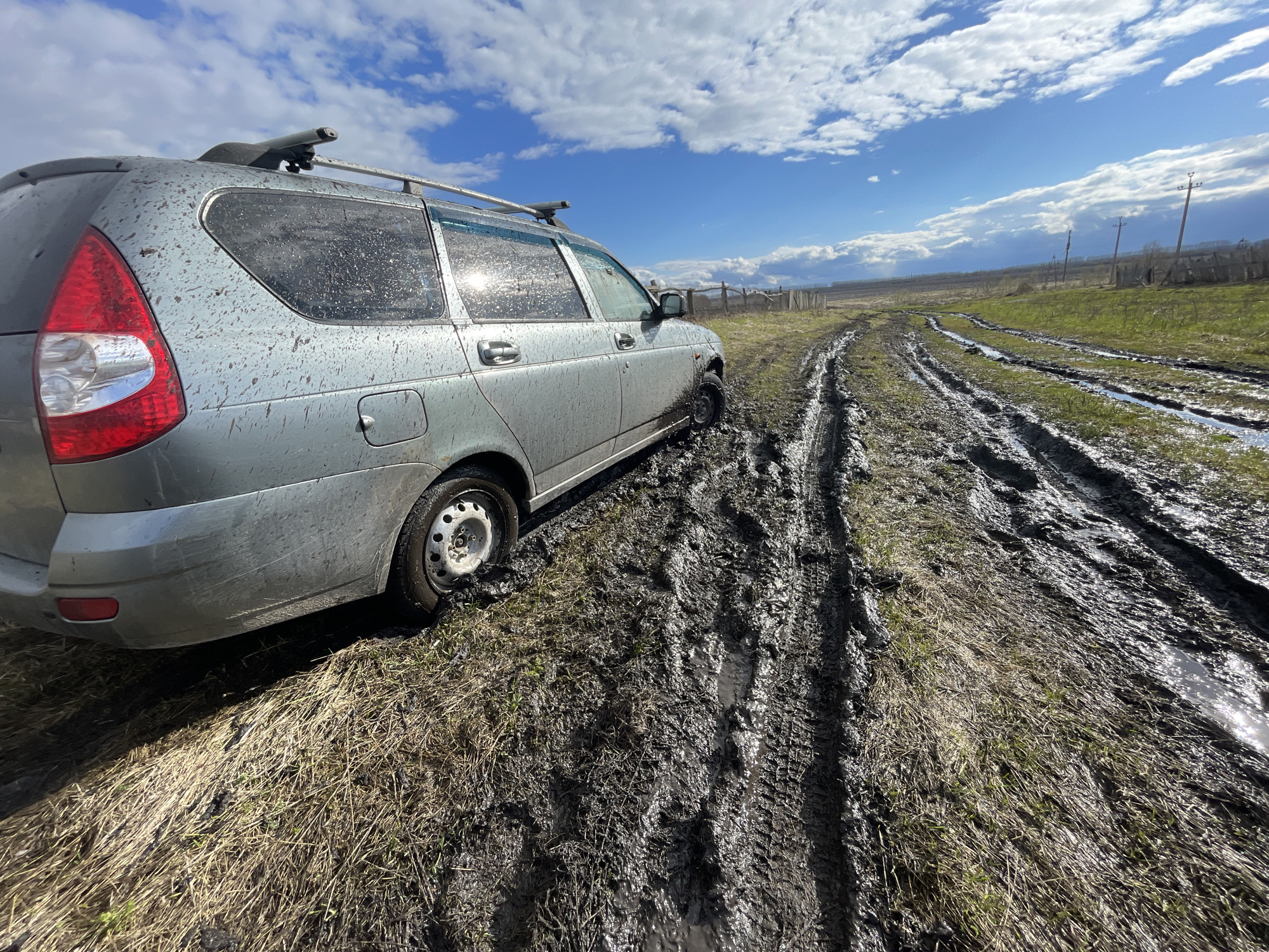 A silver Lada station wagon is stuck in a muddy dirt road in the rural area of Новая Шемурша, Russia. The car's exterior is splattered with mud. The road is deeply rutted with tire tracks filled with water. Patches of green grass and dry vegetation are visible on either side of the road. A wooden fence and telephone poles line the distant horizon under a partly cloudy sky.
