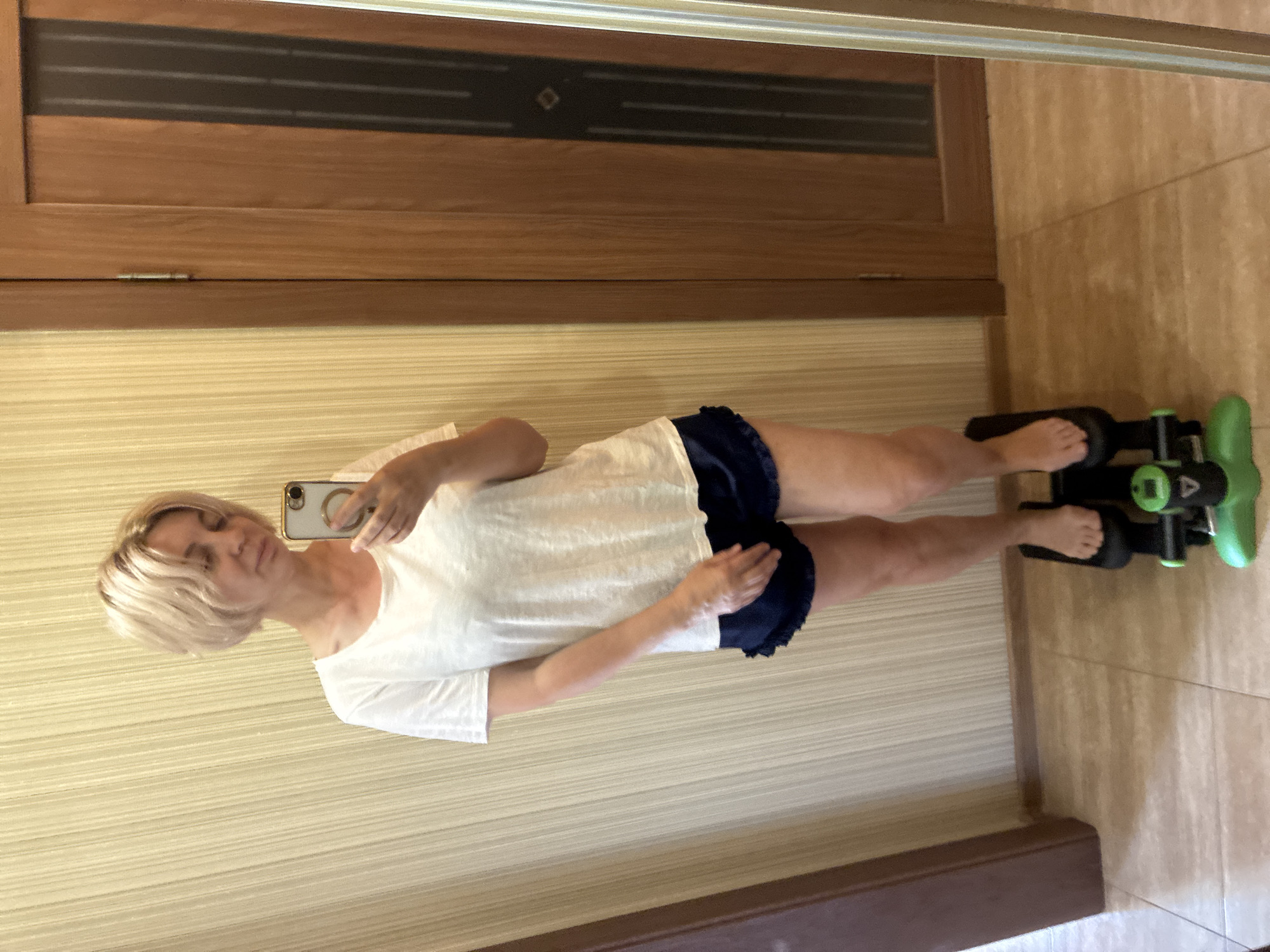 A light-skinned woman with blonde, shoulder-length hair stands on a mini-stepper indoors, holding a smartphone to take a mirror selfie. She is wearing a white t-shirt and dark blue shorts with ruffled hems. The room has light beige striped wallpaper and beige tiled flooring. To the right of the woman, a wooden door with frosted glass panels is visible. The image appears to have been taken in a residential setting.