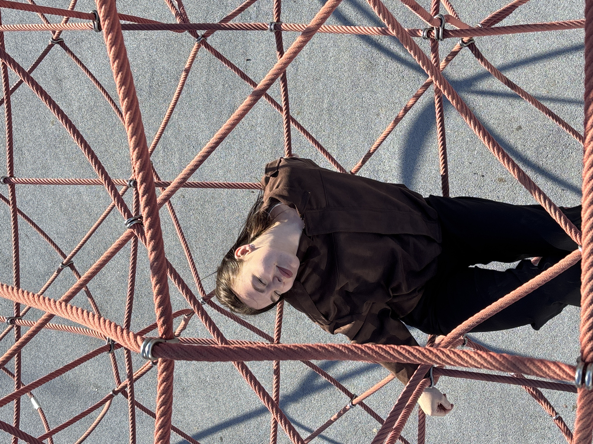 A person is lying on a rope structure in what appears to be a public space. The structure is made of reddish-brown ropes connected in a net-like pattern, suspended above a gray, textured surface. The person is wearing a dark brown jacket and black pants. The person's head is slightly tilted with their eyes closed. The location is a city, potentially Perm, Russia. The scene is captured from an overhead perspective.