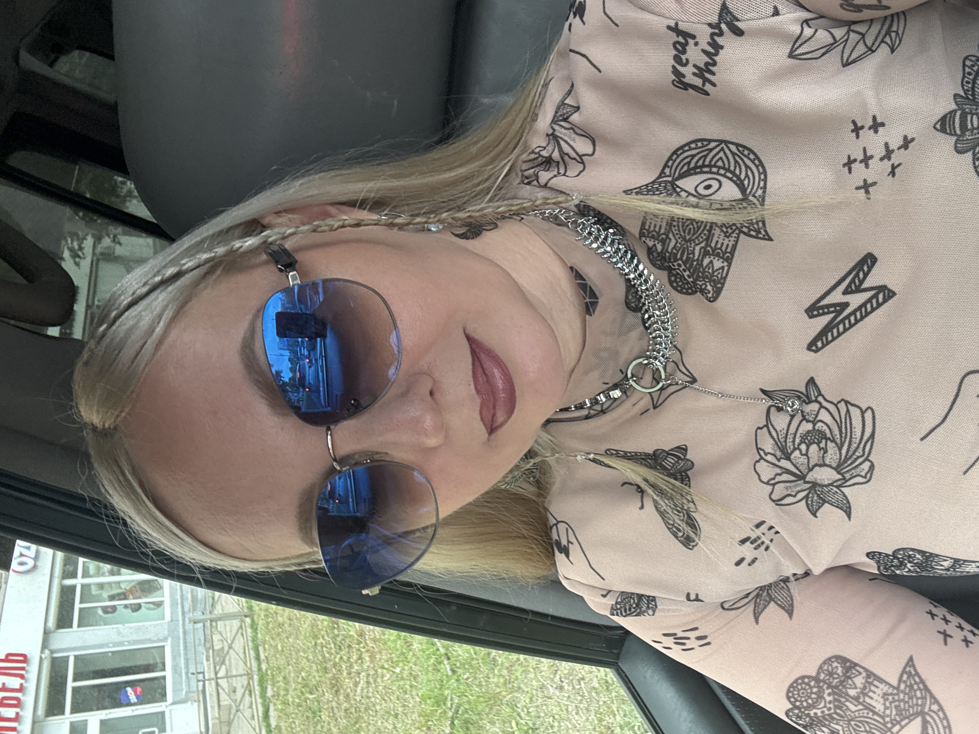 A blonde woman with blue-tinted sunglasses and braided hair is pictured inside a vehicle, likely a car. She is wearing a pink top with a dark floral and abstract pattern. A thick silver chain necklace with a ring pendant is visible around her neck. The sunglasses reflect a street scene with cars and buildings. To the left of the vehicle, a building with the Russian text "МЕБЕЛЬ" (Furniture) and "OZ" is visible, along with a grassy area. The scene appears to be in Komsomolsk-on-Amur, Russia, based on the context provided.