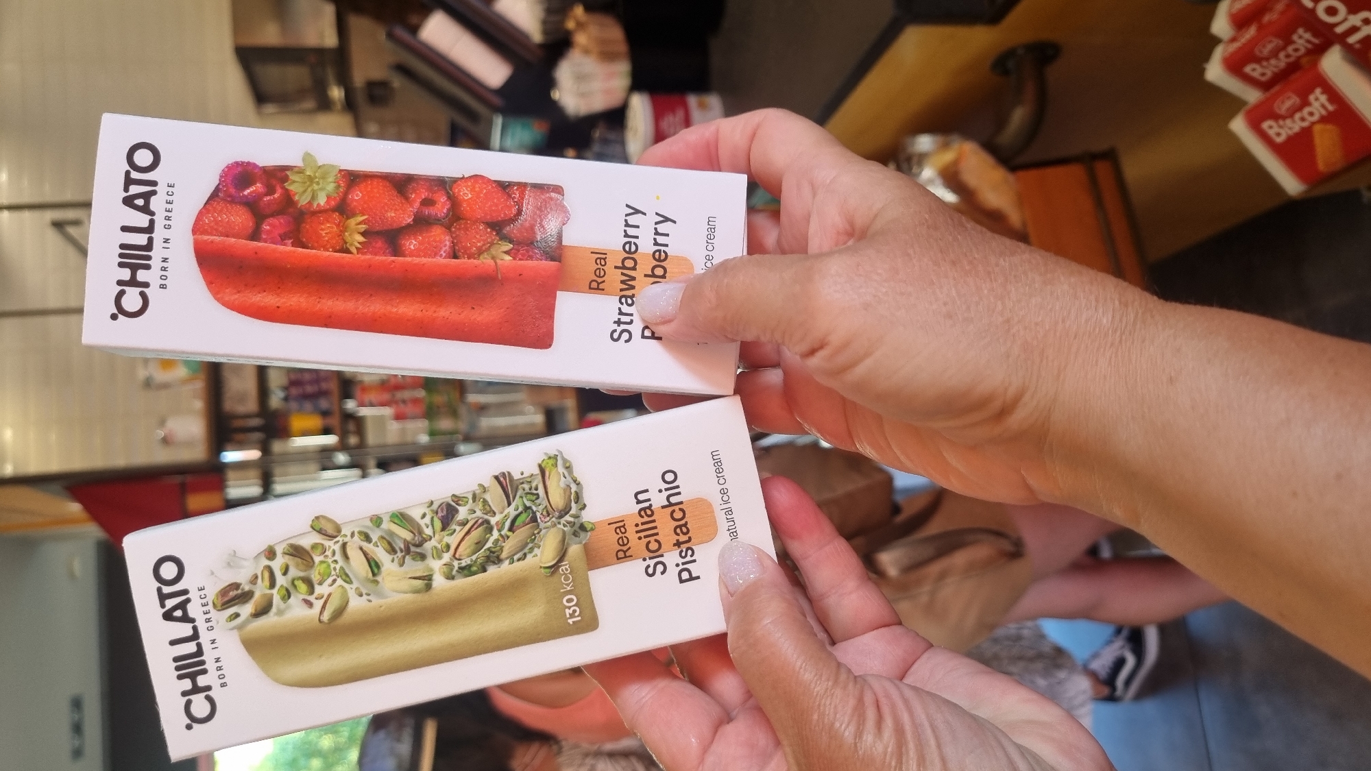In Litochoro, Greece, two hands hold up ice cream packaging. The packaging features the brand "CHILLATO" with "BORN IN GREECE" below it. The left package displays an image of pistachio nuts and a pale green ice cream bar, labeled "130 kcal" and "Real Sicilian Pistachio." The right package shows whole strawberries and raspberries and a red ice cream bar, labeled "Real Strawberry." The background appears to be an indoor setting, possibly a shop, with shelves stocked with various items, and blurred figures of people in the distance.