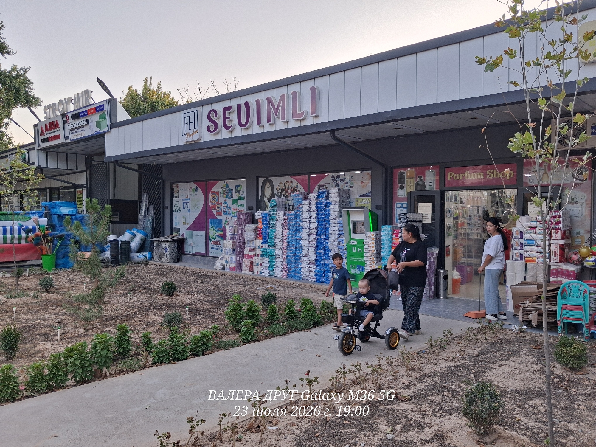 In Chirchik, Uzbekistan, on July 23, 2026, at 19:00, a family is captured on a paved walkway outside a row of shops. A woman, dressed in black, pushes a stroller containing a young child. Beside the stroller stands another child, a boy wearing a blue t-shirt. To the right, a young woman in a white t-shirt and light-colored pants sweeps the ground with an orange broom. The background features a building with several storefronts, including one labeled "SEUIMLI" and another displaying "Parfum Shop." The shops display various goods, with stacks of products visible through the windows and outside. The area in front of the shops is a mixture of dry dirt, small green bushes, and saplings. In the foreground, the walkway curves, with some wilting plants and dry earth visible along its edge. The lighting suggests it is late afternoon or early evening.