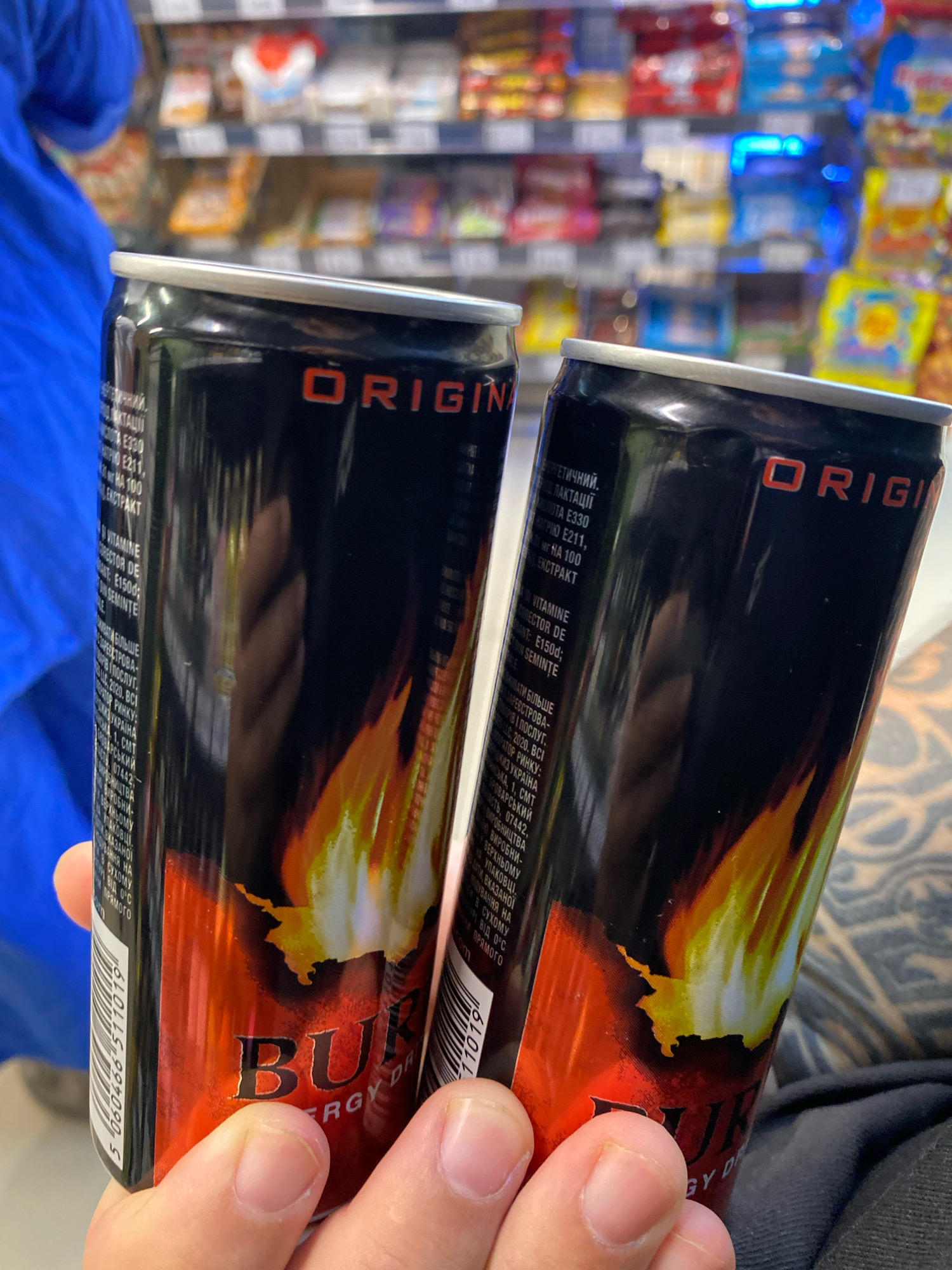 The image shows a close-up of two black aluminum cans with orange and yellow flame graphics. The cans appear to be energy drinks. Text is visible on the cans, including the word "ORIGIN" in red lettering and the partial word "BUR..." and "ENERGY DR" suggesting the brand name is "BURGER ENERGY DRINK" or similar. One can has a barcode with the number "5 060466 511019". The cans are held by a person's hands, with fingers visible. The background is blurred but appears to be a supermarket aisle with shelves stocked with various products. The lighting is artificial, consistent with an indoor retail environment. There is no indication of the location being in Chișinău, Moldova.