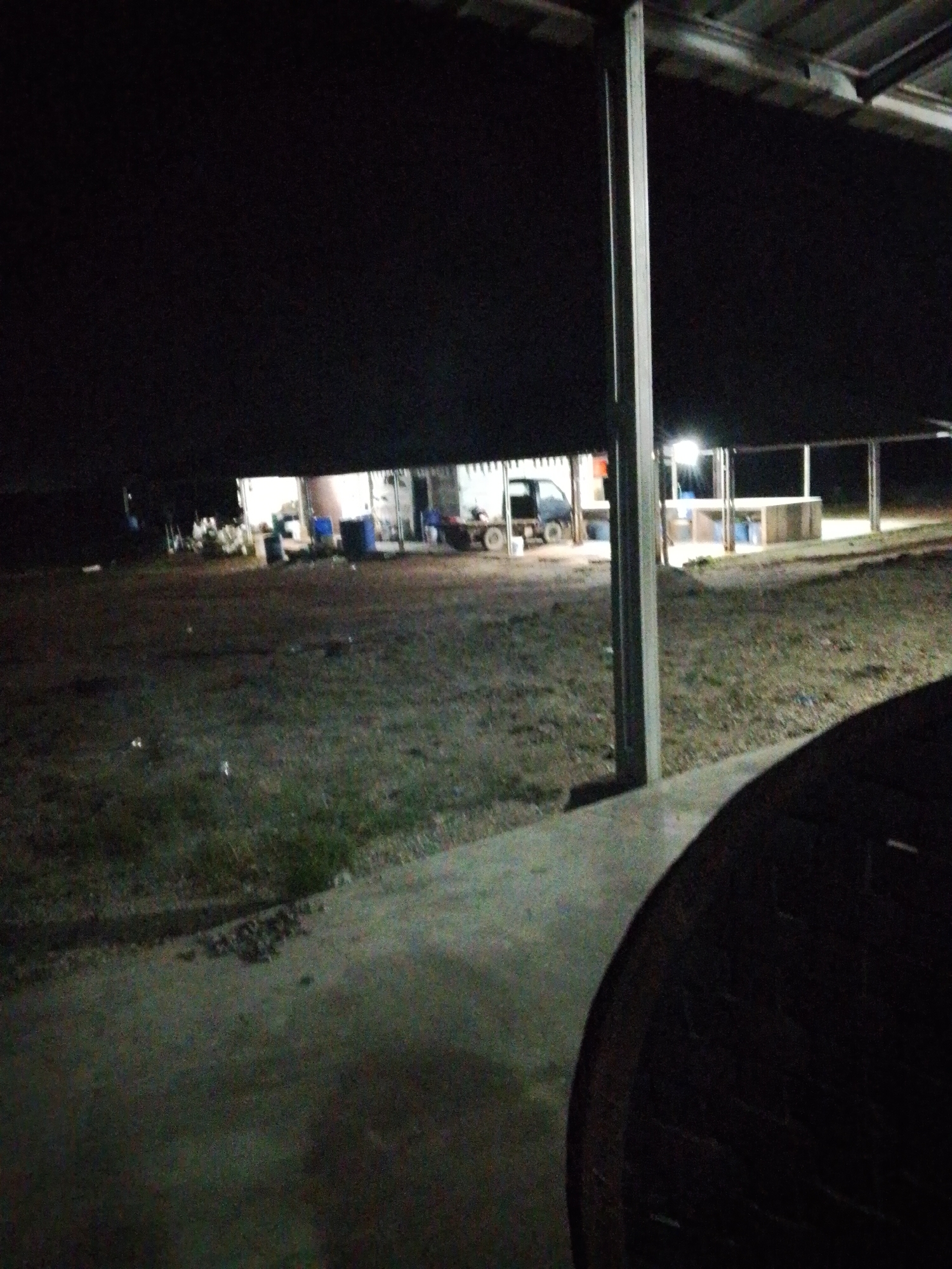 The image was taken at night in Bukit Ketok, Indonesia. It depicts an outdoor scene with a dimly lit shelter structure in the background. A small vehicle (possibly a truck) is parked near the shelter. Metal support beams are visible in the foreground. A dark, undefined object partially obscures the right side of the view. The area appears to be a yard or open space with scattered debris and vegetation.