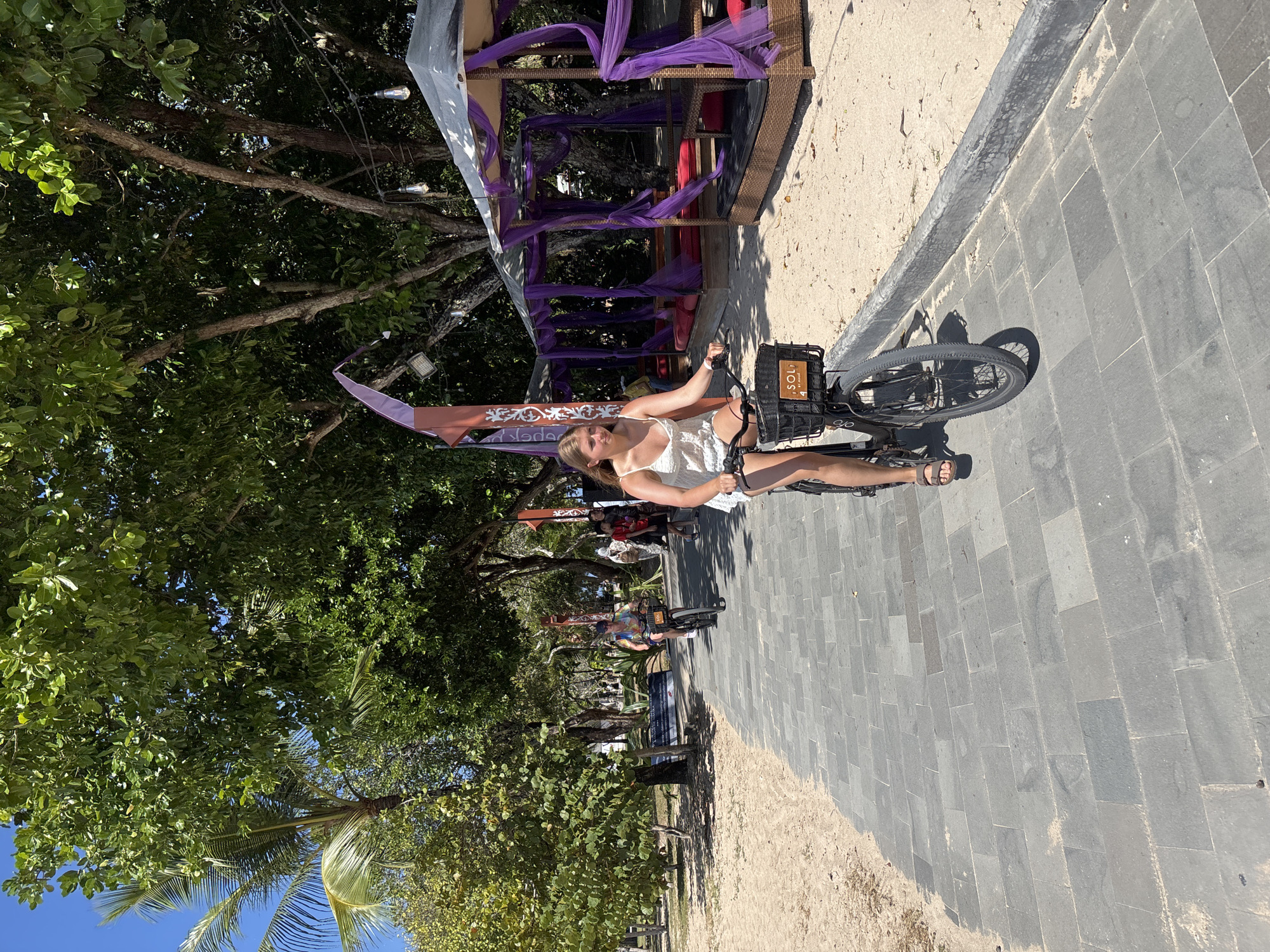 A woman in a white dress is sitting on a bicycle with a basket on the front, on a paved pathway next to a sandy beach. She is looking towards the camera with a smile. Behind her is a shaded structure with purple fabric draped over it. Trees with lush green leaves and palm fronds line the area, with a clear blue sky visible through the branches. The scene appears to be outdoors during the daytime, likely on a sunny day given the shadows cast by the bicycle. The woman's attire and the beach setting suggest a tropical or coastal location. There is text visible on a sign behind the woman and on the basket of the bicycle. The sign behind her appears to have decorative patterns and some writing that is not fully legible. The basket has a label that says "SOL". The overall mood is relaxed and pleasant.