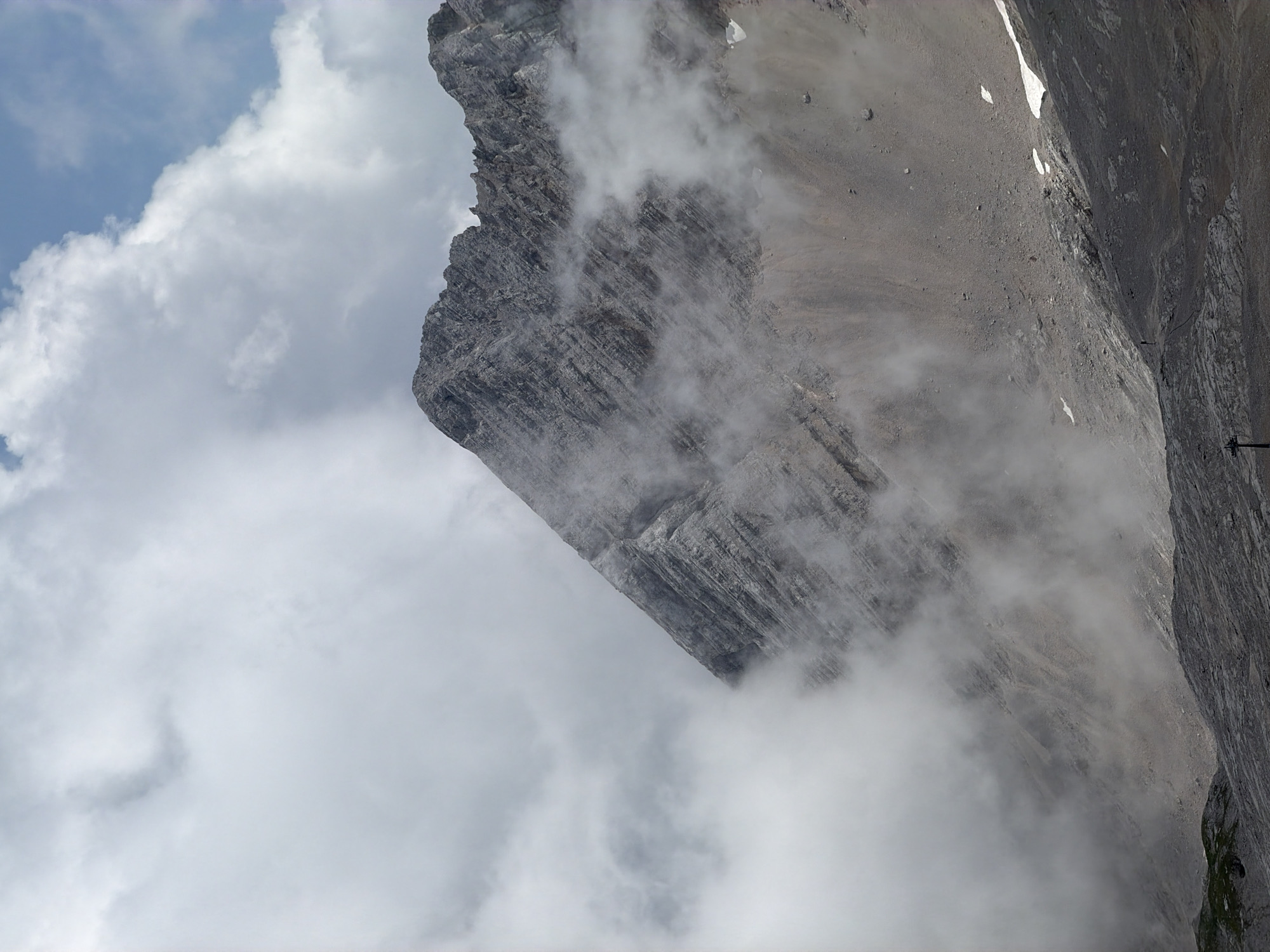 The image depicts a dramatic, overcast mountain landscape with rocky, striated slopes partially obscured by swirling mist. The prominent mountain peak in the center is textured with visible layers of rock and appears steep and imposing. Patches of white snow are visible on some of the lower slopes. In the foreground, a rugged, dark, rocky terrain stretches out, suggesting a high-altitude environment. A solitary, small human figure can be observed standing on the lower slope, silhouetted against the landscape, indicating a scale comparison. The sky above is a mix of bright white clouds and patches of blue. This scene is consistent with the Zugspitze, Germany's highest peak, located in the Bavarian Alps near Garmisch-Partenkirchen.