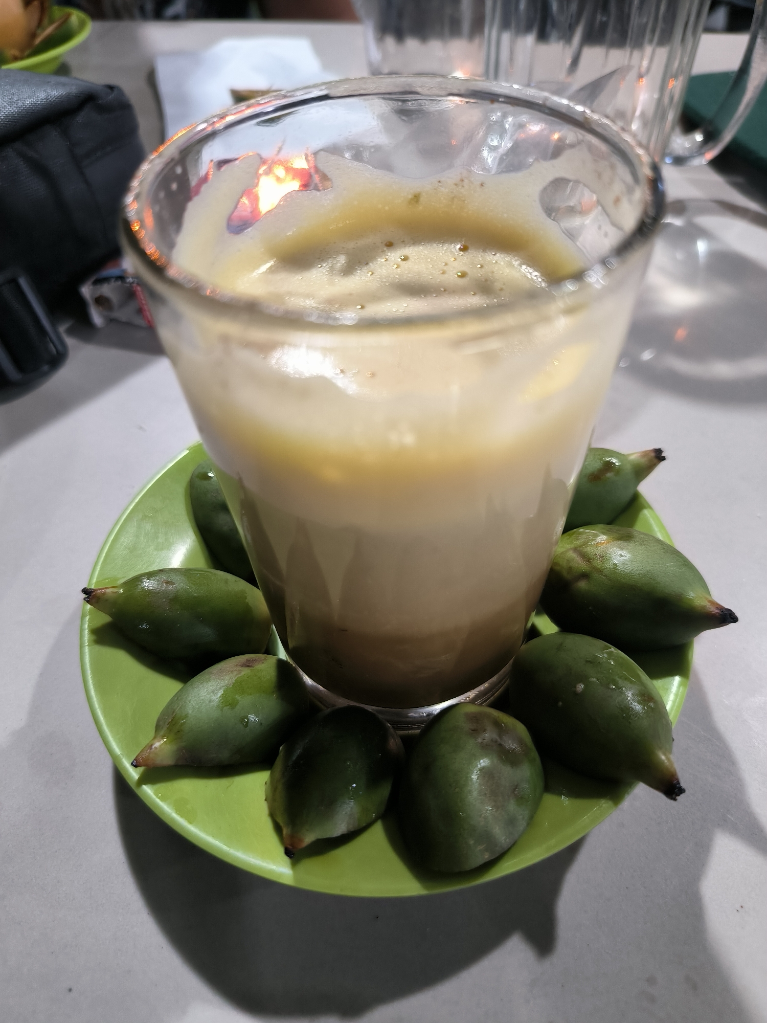 A glass of light brown beverage rests on a small green plate. The beverage has a foamy top and appears layered. Several small, green, teardrop-shaped fruits with dark tips encircle the base of the glass on the plate. The plate sits on a light gray surface. In the background, portions of a dark-colored bag, a clear pitcher, and other items are visible, but out of focus. The lighting suggests an indoor setting with artificial illumination. Given the context of Lhokseumawe, Indonesia, the green fruits may be "buah nipah" (Nypa palm fruits), which are sometimes served alongside beverages.