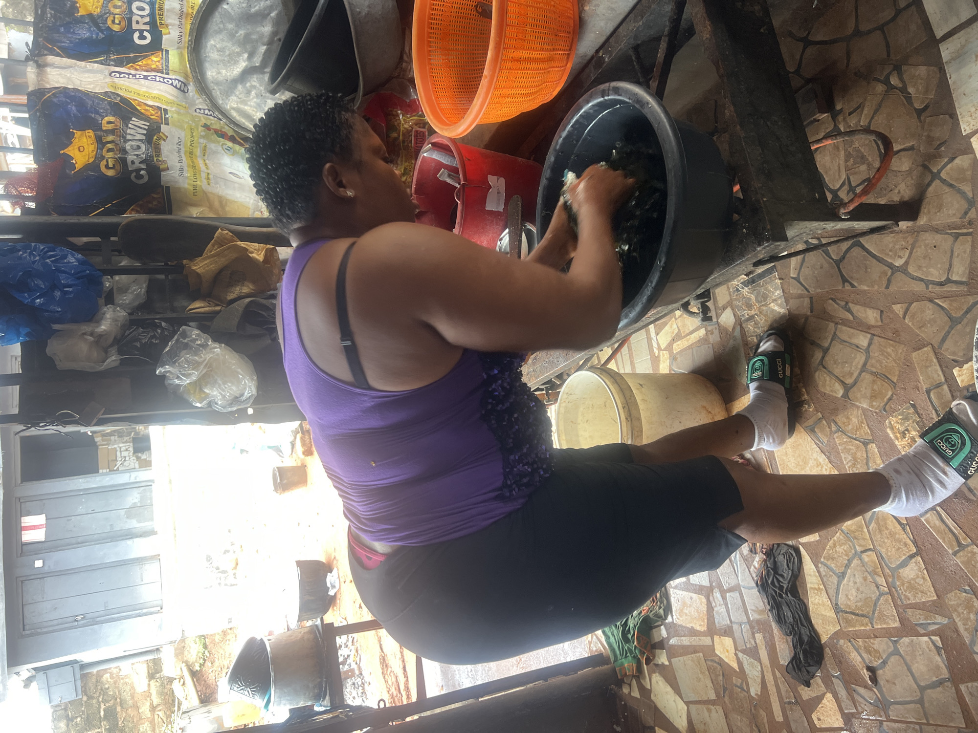 In Nkpor, Nigeria, a woman is preparing food in what appears to be an outdoor market stall. She is wearing a purple tank top and black skirt, and is crouched over a large black basin filled with water and leafy greens. She is actively washing the greens with her hands. The setting includes a metal stove, various containers, and bags of rice in the background. The ground is tiled and appears wet.