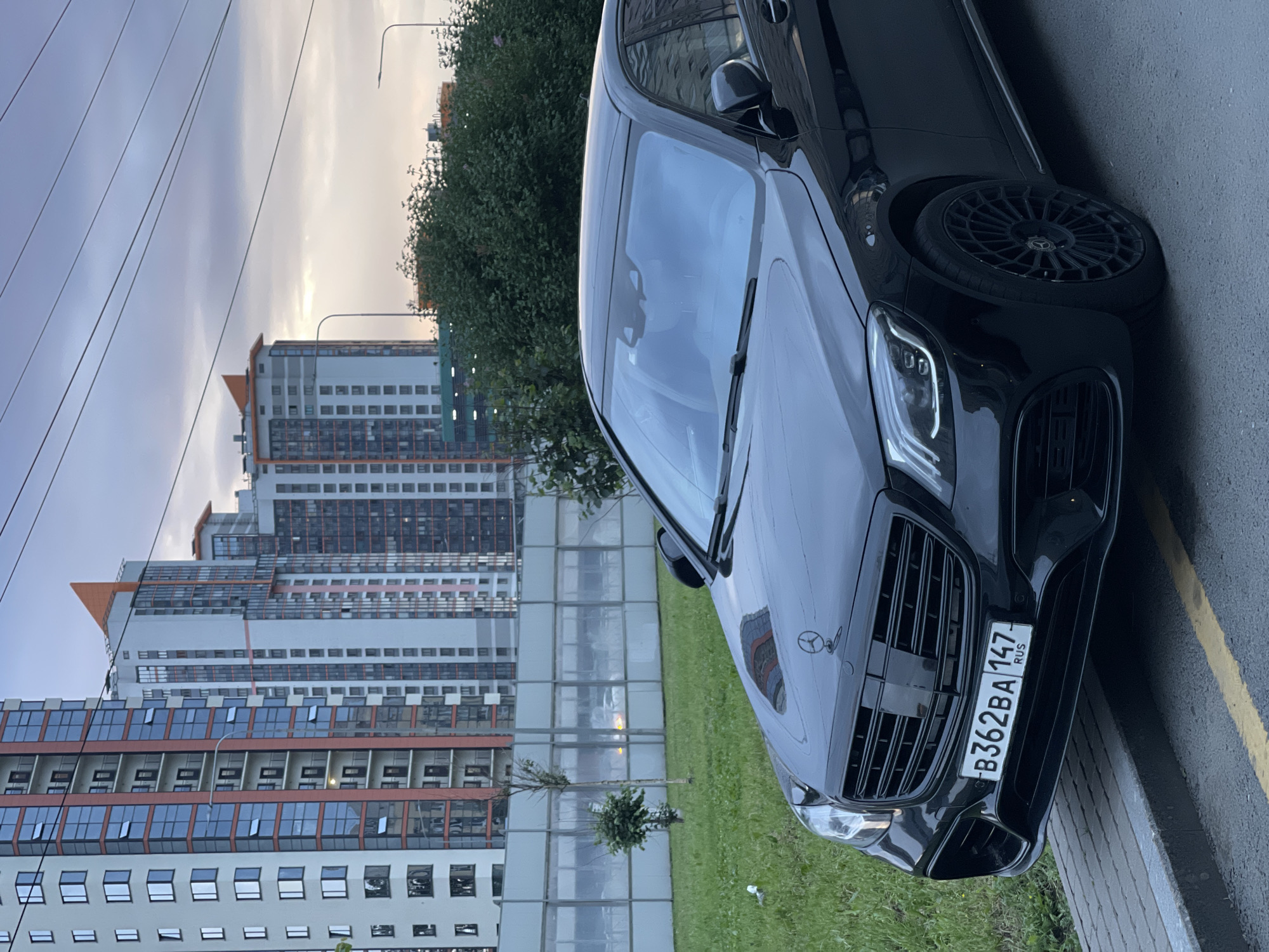 A black Mercedes-Benz S-Class sedan is parked on a street in front of several modern high-rise buildings and a row of trees. The car's front end, including the grille, headlights, and license plate which reads "B362BA 147 RUS," is prominently visible. The sky is cloudy, with hints of the sunset visible. The buildings in the background are tall and appear to be residential or commercial, with a mix of white, grey, and orange facades, and numerous windows. A short, thin tree and a small shrub are located on the grassy median between the sidewalk and a transparent fence. The street appears to be paved with asphalt, and a yellow line is visible on the road surface.