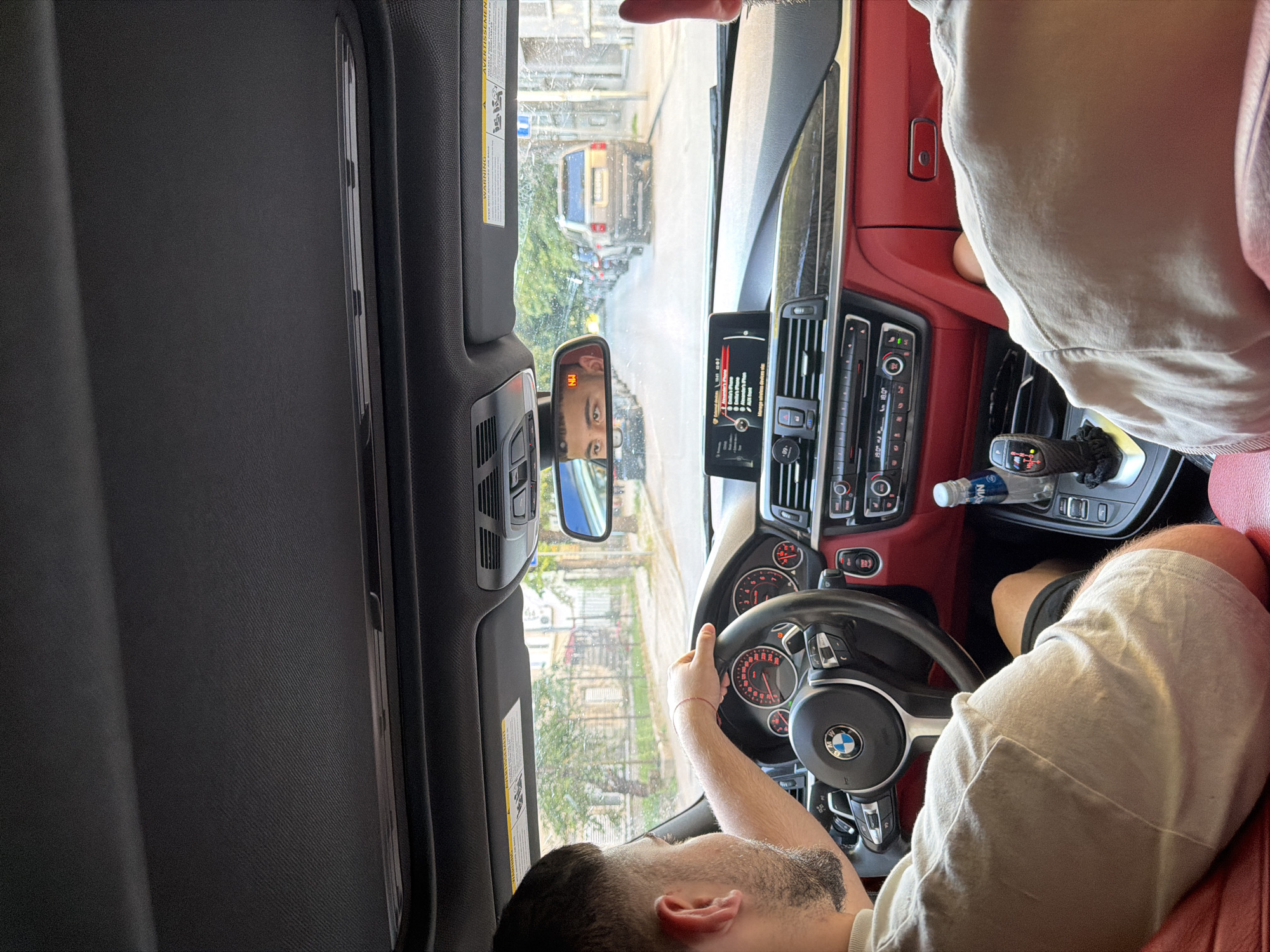 The image shows the interior of a car, likely a BMW based on the steering wheel emblem, with a man driving. He is wearing a light-colored t-shirt and dark shorts, and his left hand is on the steering wheel. The car's dashboard is visible, featuring a modern infotainment system and red accents on the interior panels. Outside the car, through the windshield and side windows, a street scene is depicted with buildings and trees. A side-view mirror reflects the driver's face and the road behind the car. The presence of a water bottle in the center console and a red string bracelet on the driver's wrist are notable details. The overall impression is a snapshot of everyday life in a city, with the specific street and buildings suggesting a location in Ruse, Bulgaria, though this cannot be definitively confirmed from the image alone.
