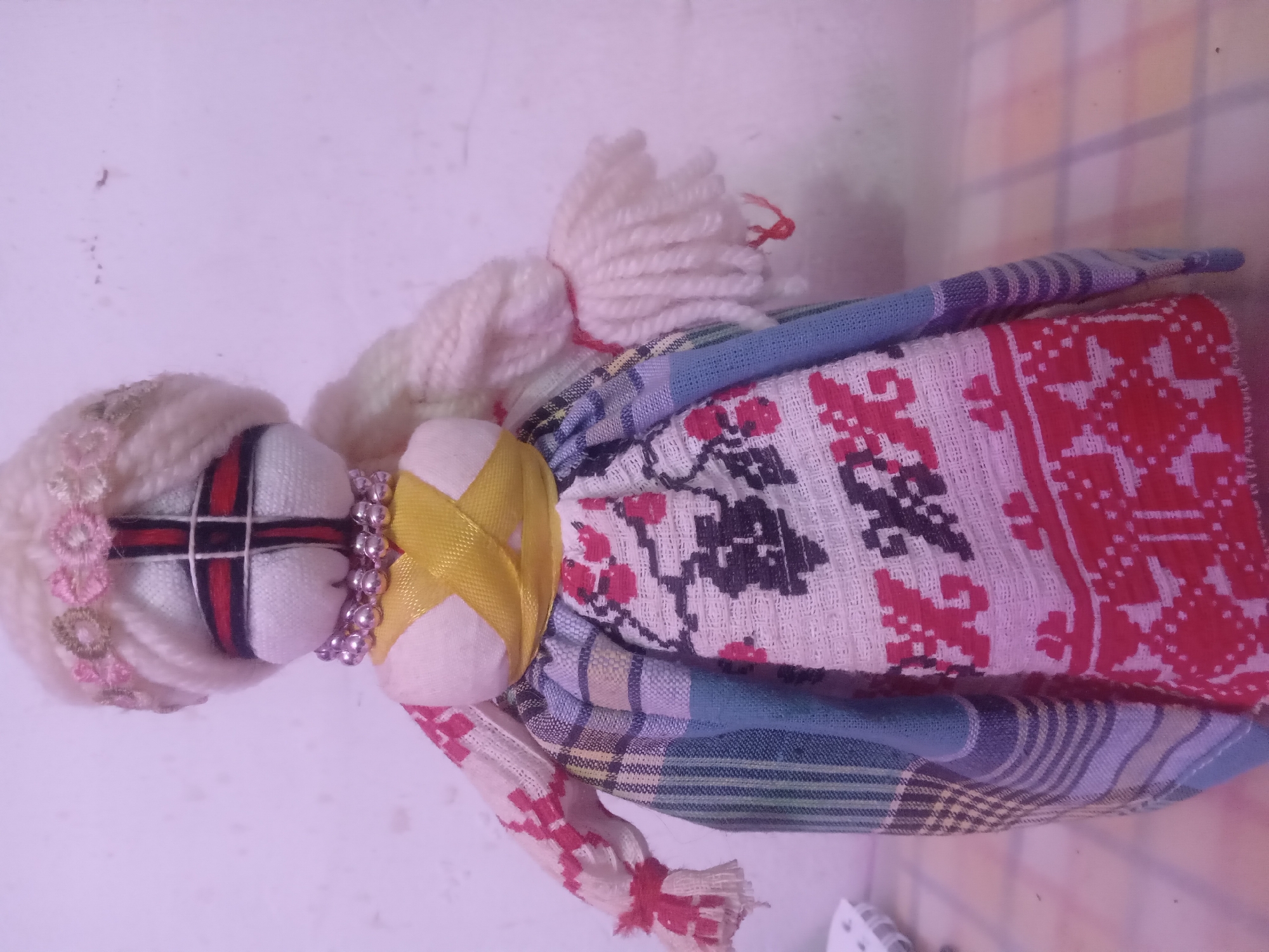 The scene captures a handmade folk art doll within an indoor environment in Unknown, Unknown. The doll, crafted from fabric and yarn, stands upright, centered in the frame. Its head is a white fabric sphere, devoid of distinct facial features, instead displaying a prominent black vertical line intersected by a red horizontal line, forming a cross. Thick, off-white yarn forms the doll's hair, styled into two long braids or plaits extending down each side. A decorative band of small, light pink and gold floral elements adorns the top of the doll's head. Around its neck, a necklace composed of silver-colored spherical beads is visible. The doll's torso is made of white fabric, with a yellow ribbon tied in a criss-cross pattern across the chest and cinching the waist. The arms, also made of white fabric, feature sleeves with a red and white patterned design, terminating in frayed, fringed cuffs secured by red thread. The arms are gently bent. The doll wears a full, multi-panelled skirt. The central front panel is white with a repeating pattern of red and black geometric and stylized floral motifs. Adjacent panels include sections with vertical blue and white stripes, and other sections with broader plaid or checkered patterns in shades of blue, green, and gray. The doll is positioned against a plain, light-colored wall that exhibits minor smudges and dust specks. It rests on a subtle light-colored, patterned surface in the foreground. The ambient lighting gives the scene a slight purplish cast.