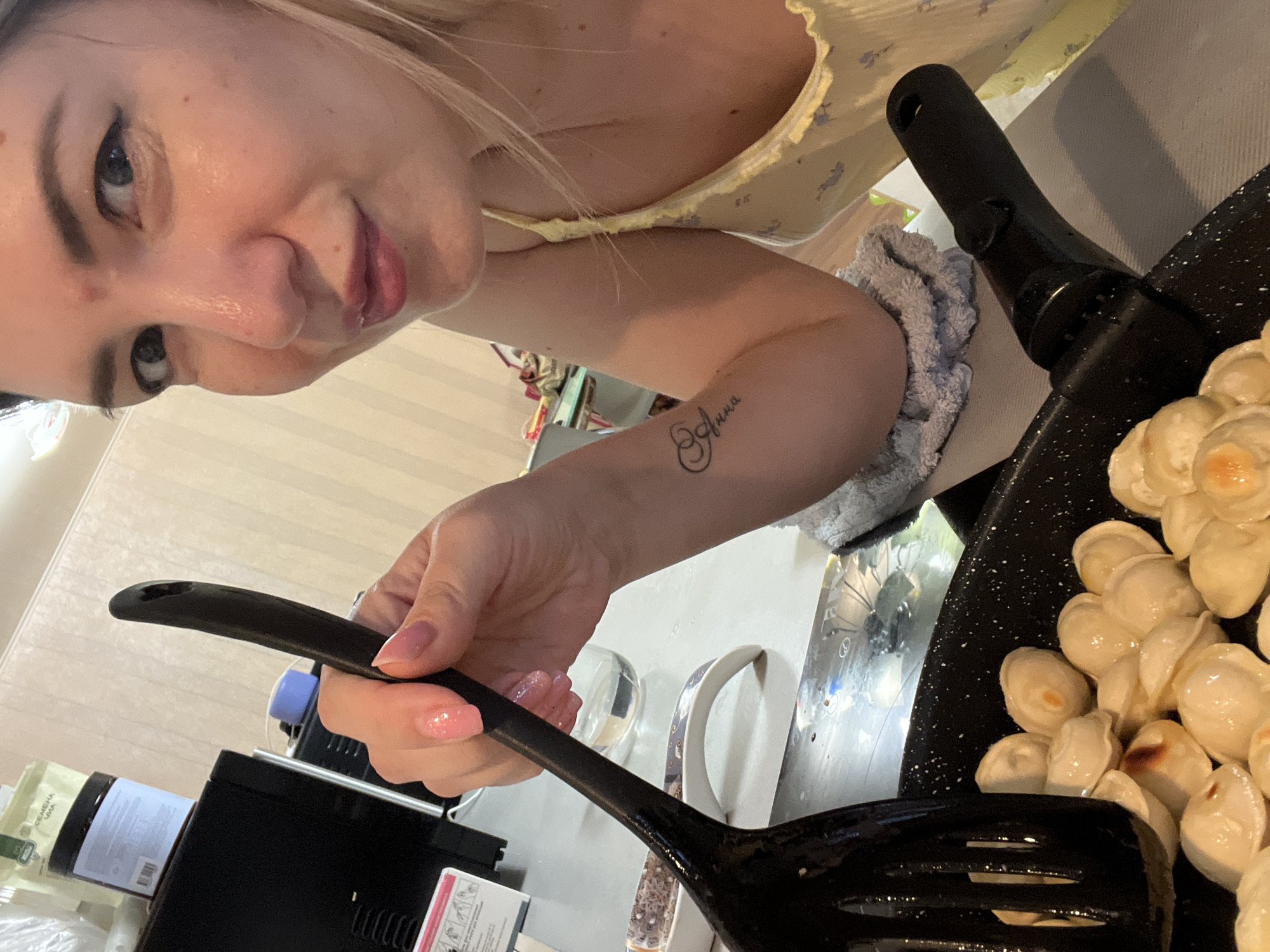 A close-up, high-angle shot captures a woman in her 20s with blonde hair and fair skin, looking directly at the camera with a playful expression. She is holding a black slotted spatula over a frying pan filled with pelmeni, a type of dumpling, which appear to be frying or boiling. The woman is wearing a yellow floral top. A tattoo of the name "Anna" is visible on her left forearm. The scene takes place in an indoor setting, likely a kitchen, with a microwave and other kitchen items visible in the background. The lighting is bright, creating a warm atmosphere. There is no indication of the specific location within Sochi, Russia.